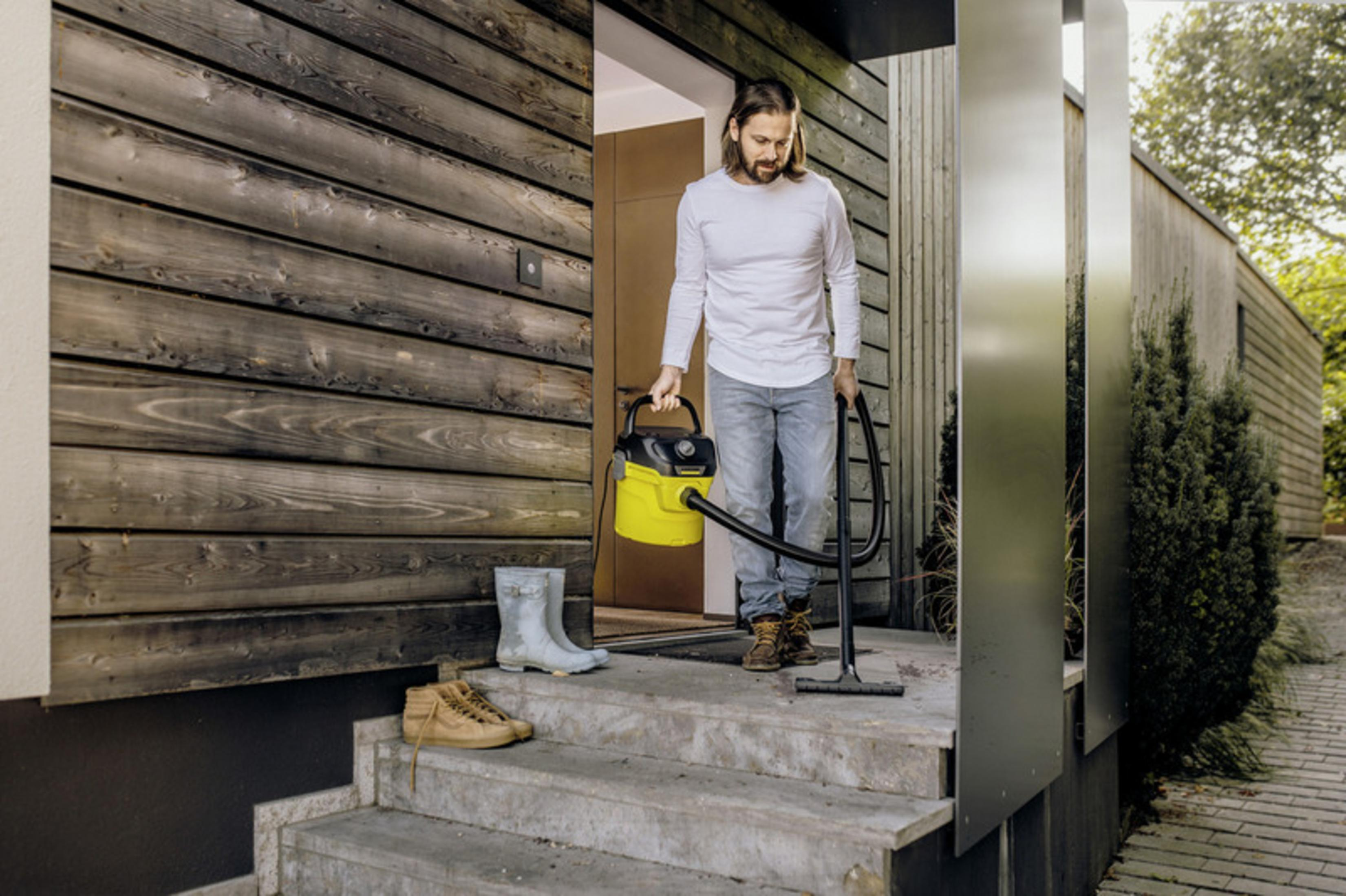 Ein Mann mit langem Haar, weißem Shirt und Jeans verlässt ein Holzhaus und trägt einen gelben und schwarzen Staubsauger.