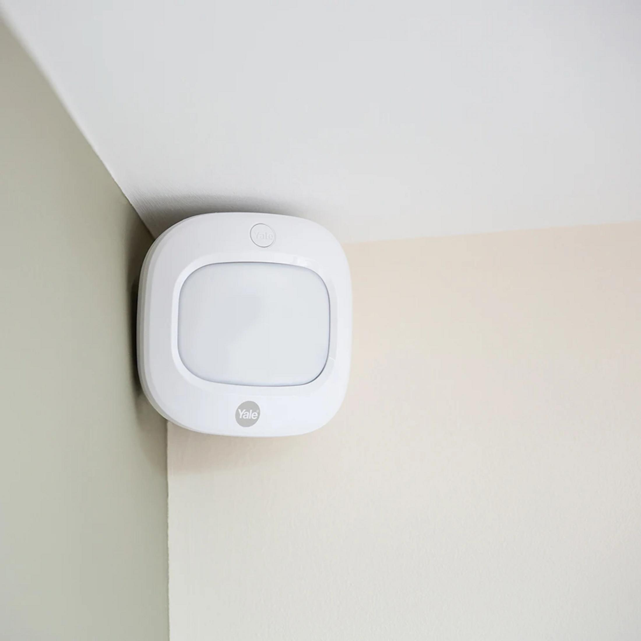 Ein weißer Yale Smart-Sicherheitsensor ist in der Ecke eines Raumes an beige-weißen Wänden angebracht.
