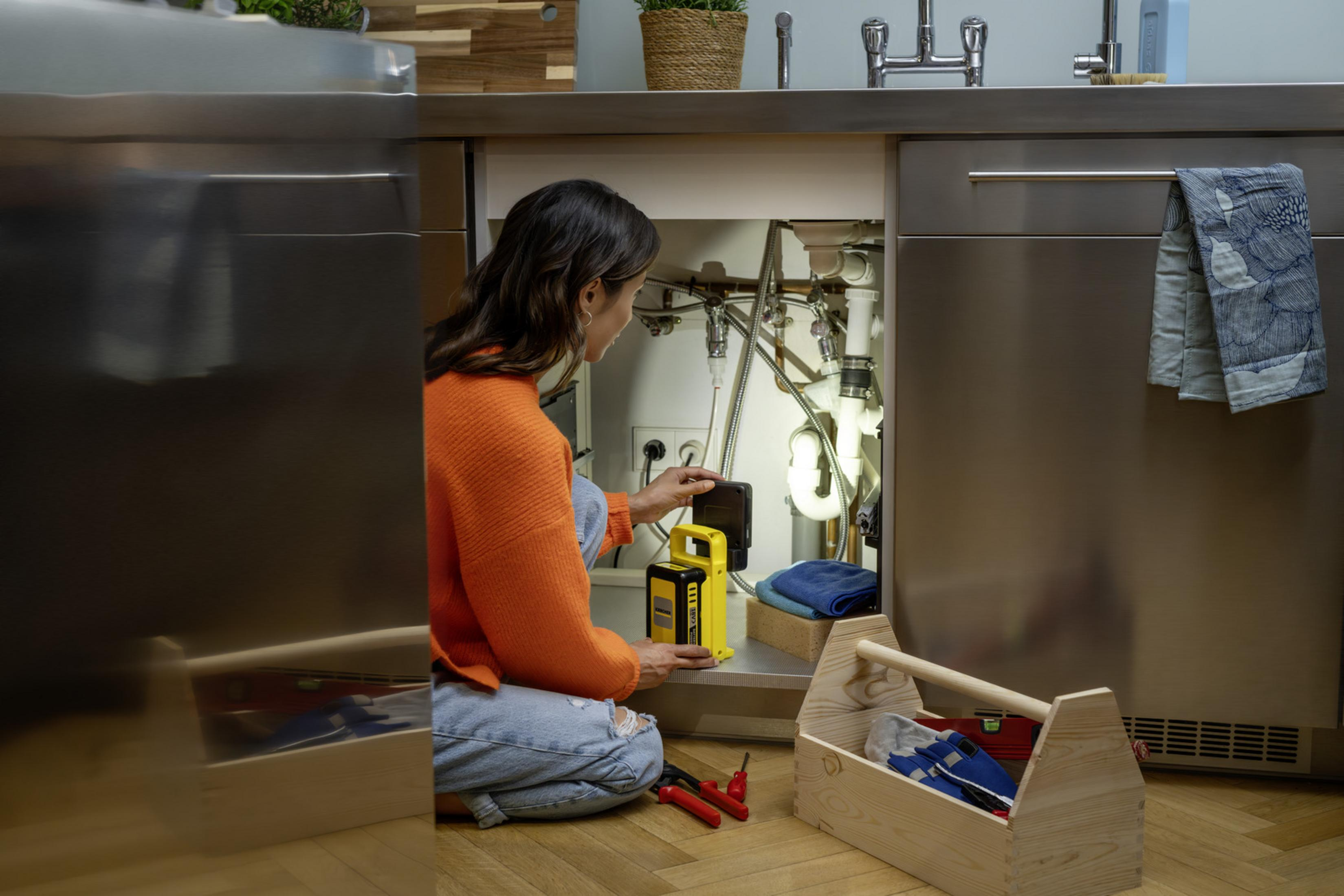 Una mujer con un suéter naranja se arrodilla ante un mueble de cocina, iluminando las tuberías.