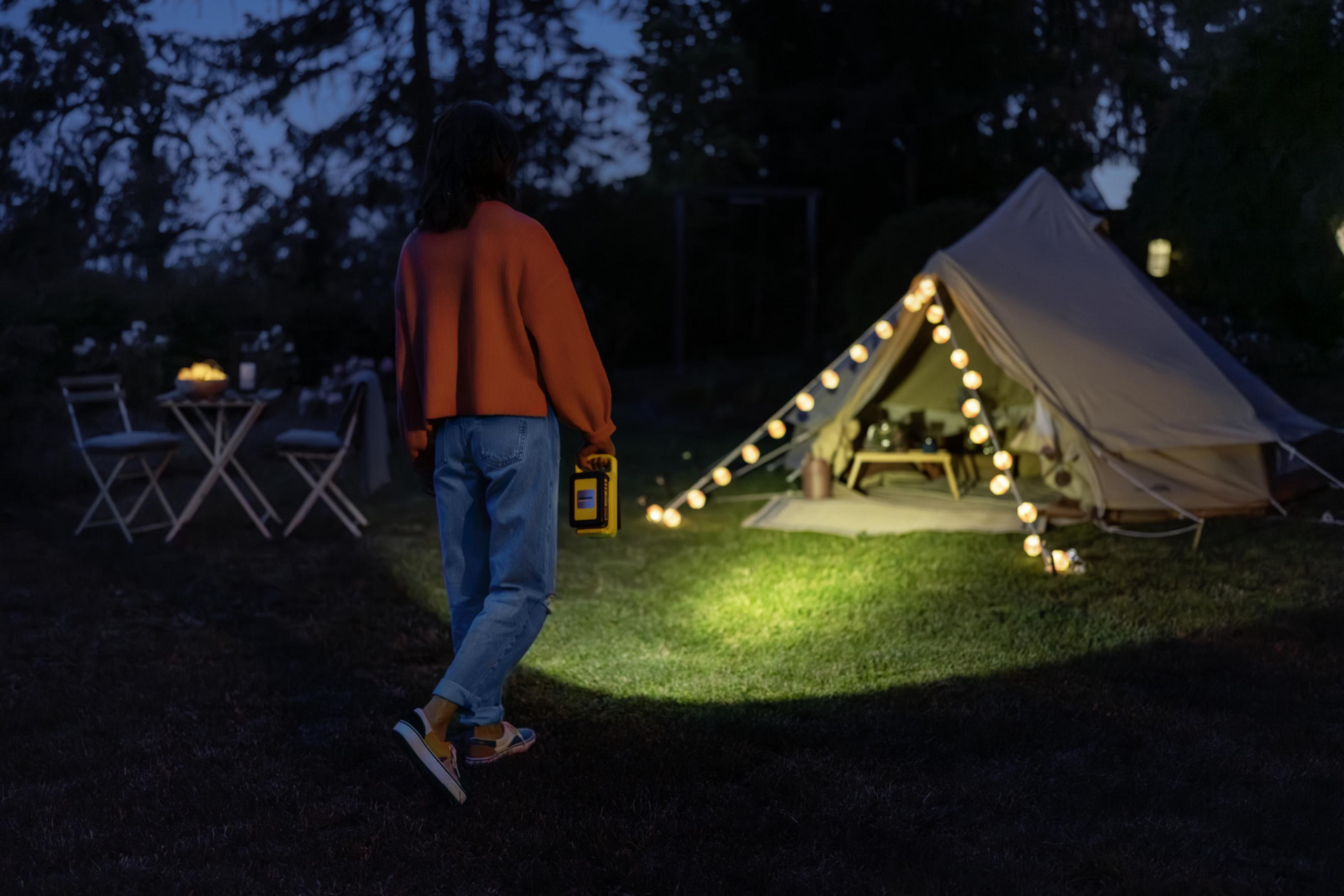 Una mujer sostiene un farol en un campamento al atardecer, con una tienda iluminada.
