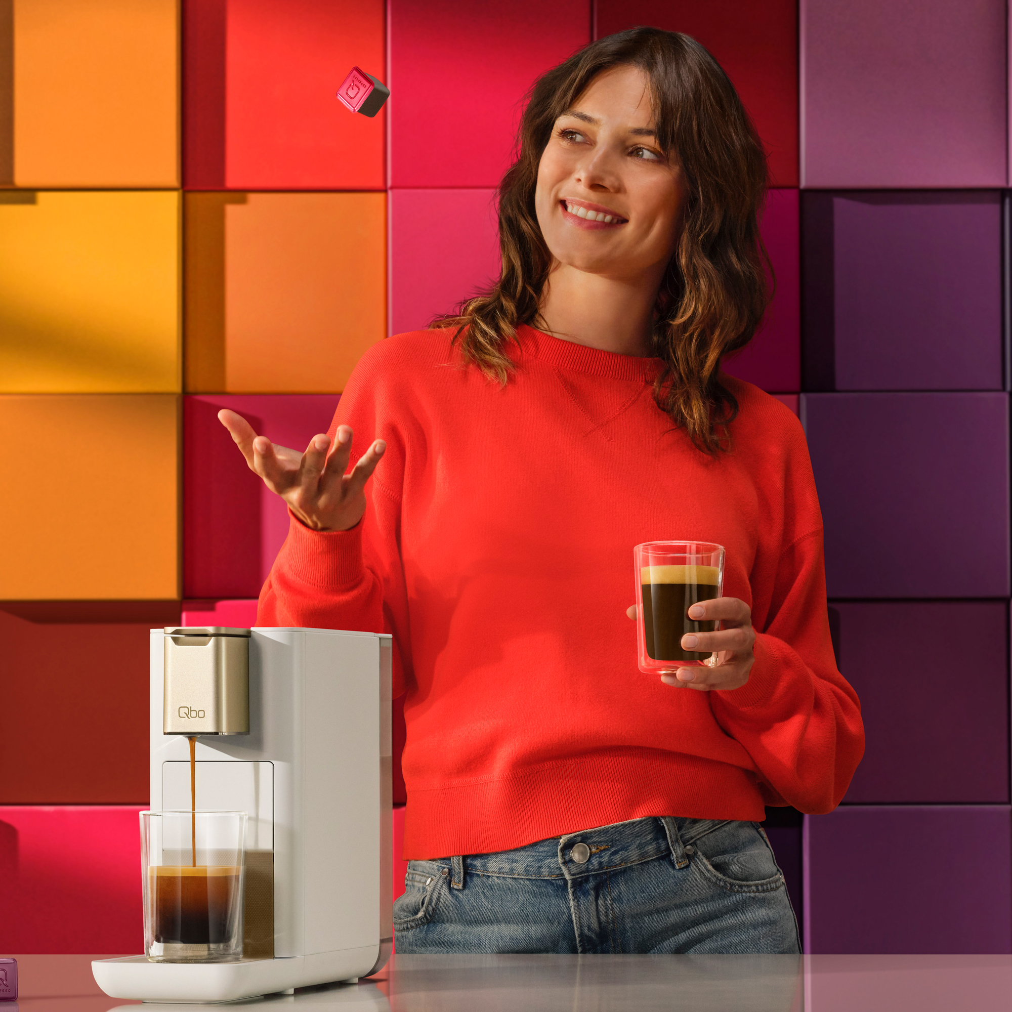 Frau in rotem Pullover hält Kaffeeglas, während eine Kapsel in die Luft fliegt, neben einer Kaffeemaschine.