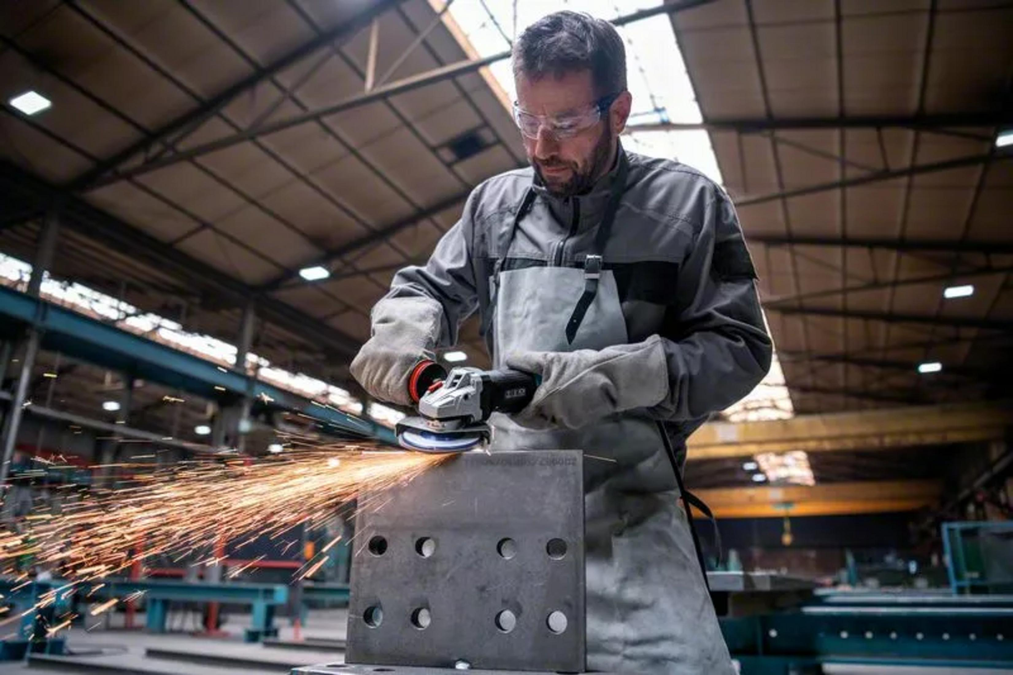 Ein Arbeiter in Schutzbrille und Handschuhen nutzt eine Flex, während Funken in einer großen Fabrikhalle sprühen.