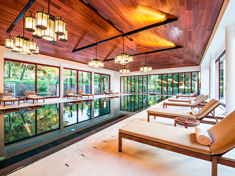 Lujosa piscina cubierta con techo de madera, grandes ventanas, tumbonas y lámparas cálidas.