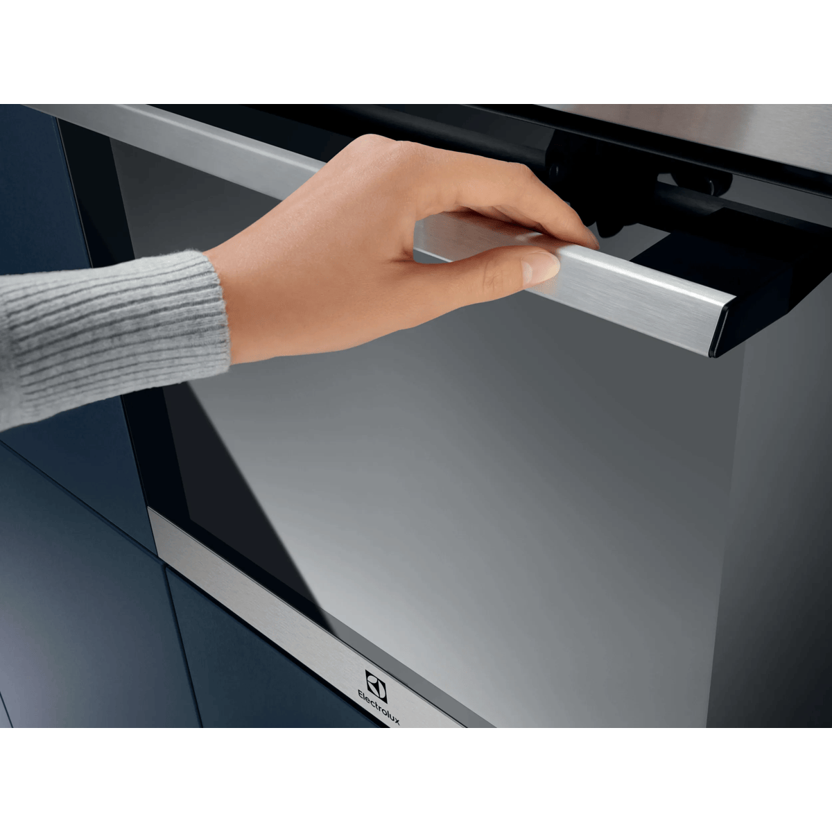 Una mano abre el tirador plateado de un horno Electrolux moderno en una cocina azul oscura.