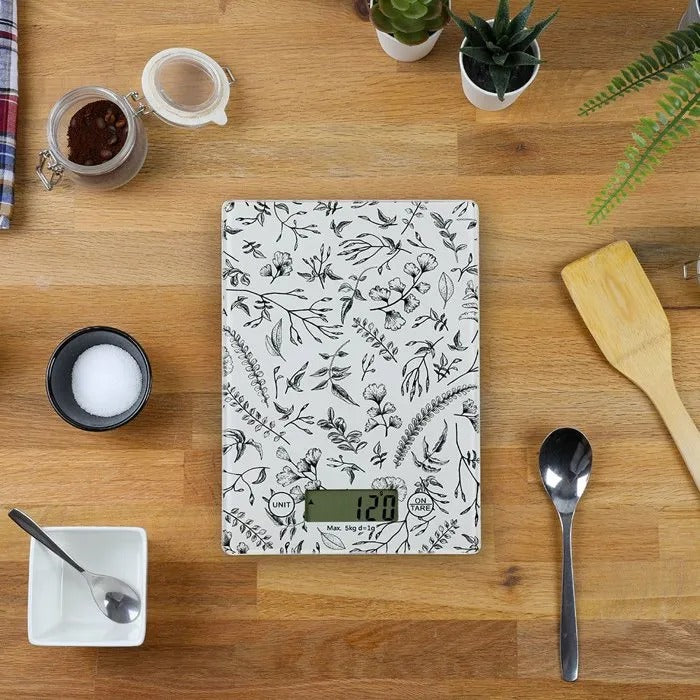 Una balanza de cocina floral con cuchara, frasco y espátula de madera sobre una mesa.