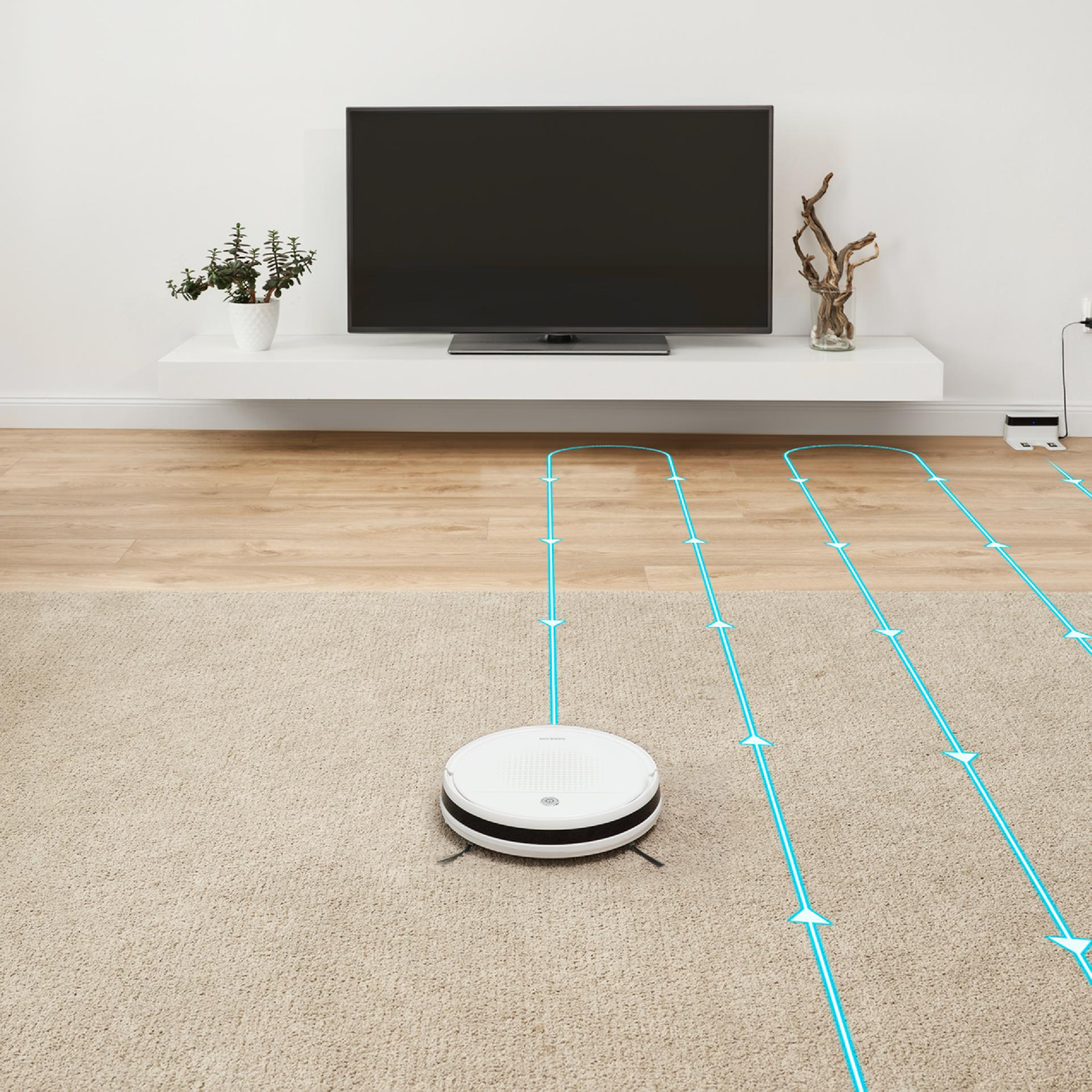 Un robot aspirador blanco limpia la alfombra en una habitación con suelo de madera y TV.