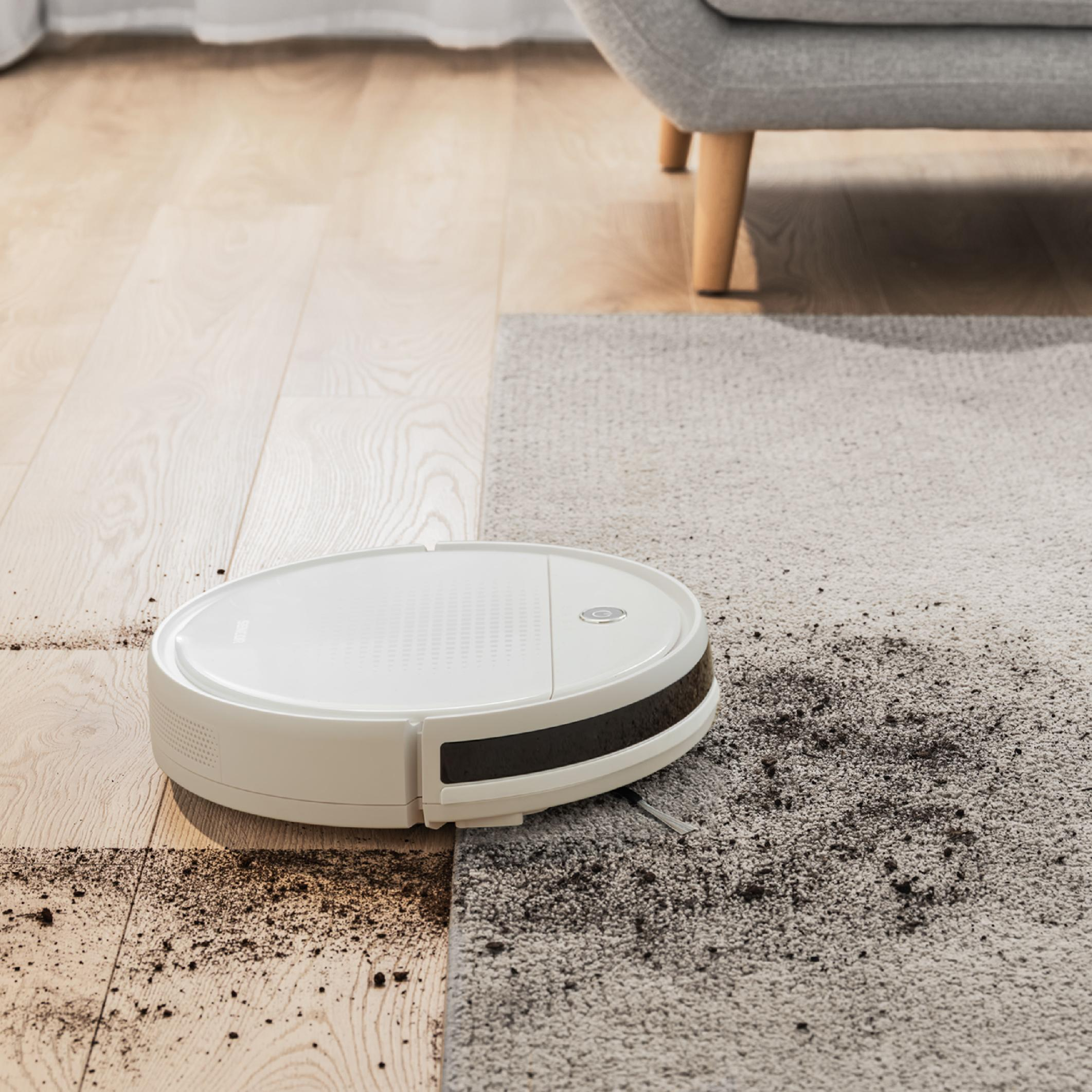 Un robot aspirador blanco limpia la suciedad de un suelo de madera y alfombra.