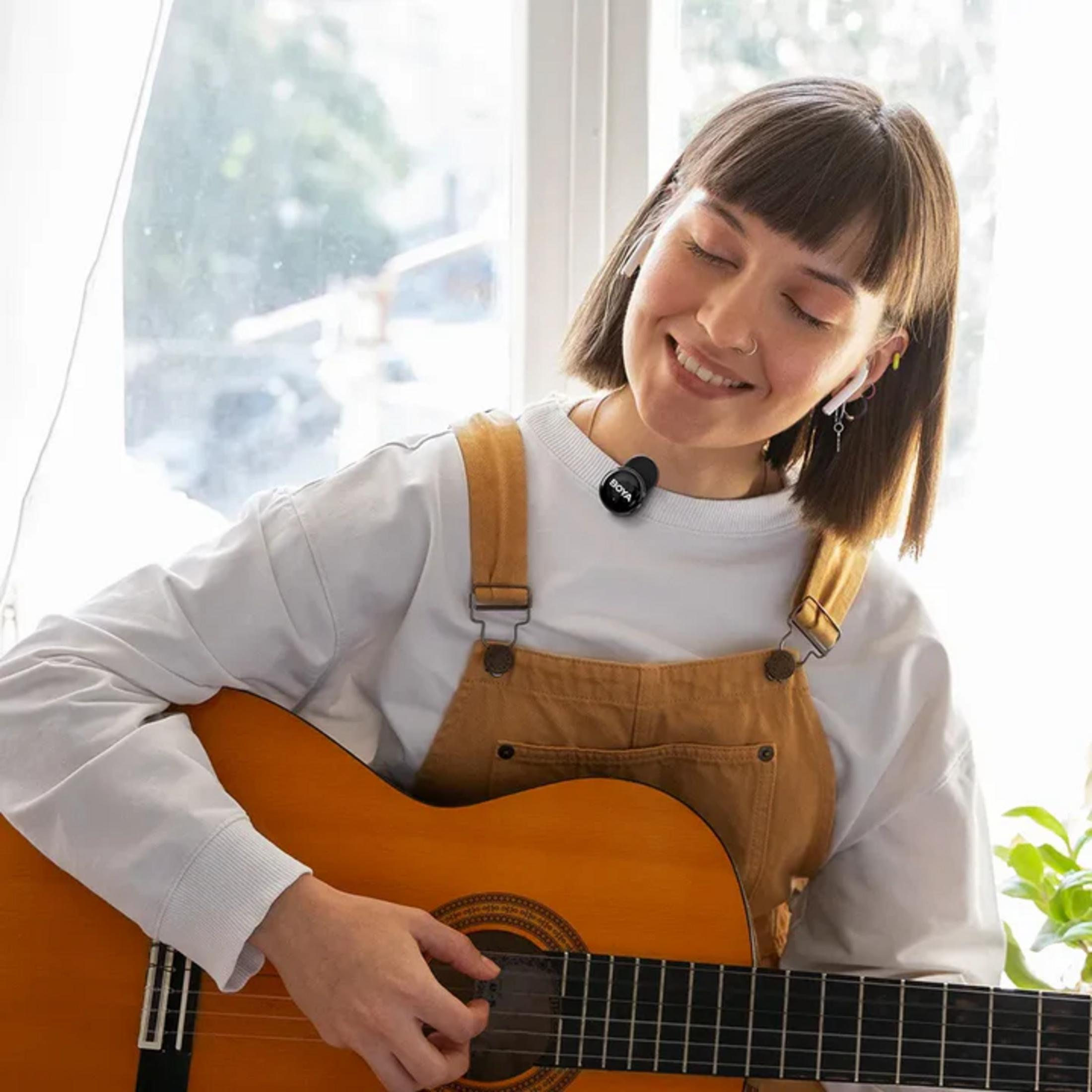 Kısa kahverengi saçlı ve tulum giyen bir kadın, küçük bir mikrofonla neşeyle gitar çalıyor.