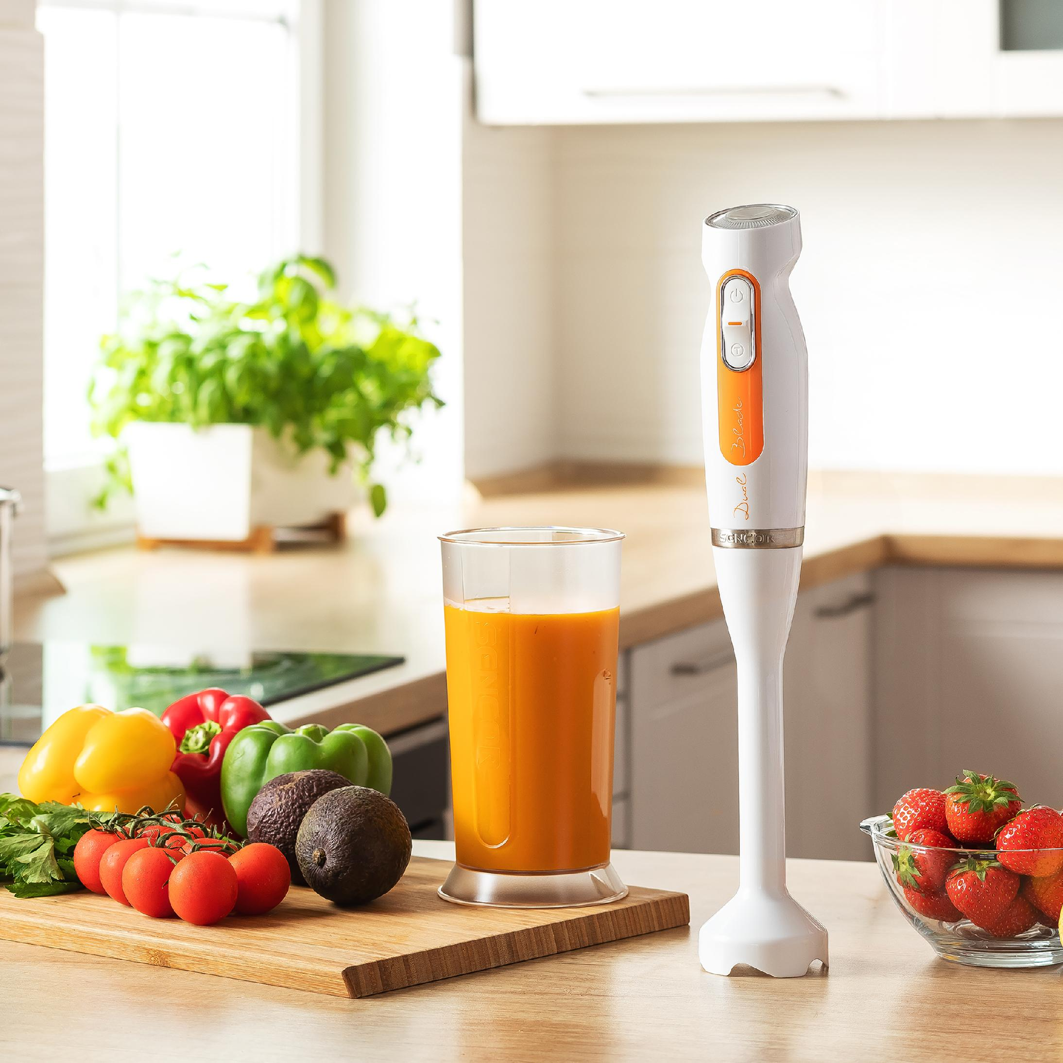 Biały blender ręczny stoi na blacie kuchennym obok świeżych warzyw, smoothie i truskawek.