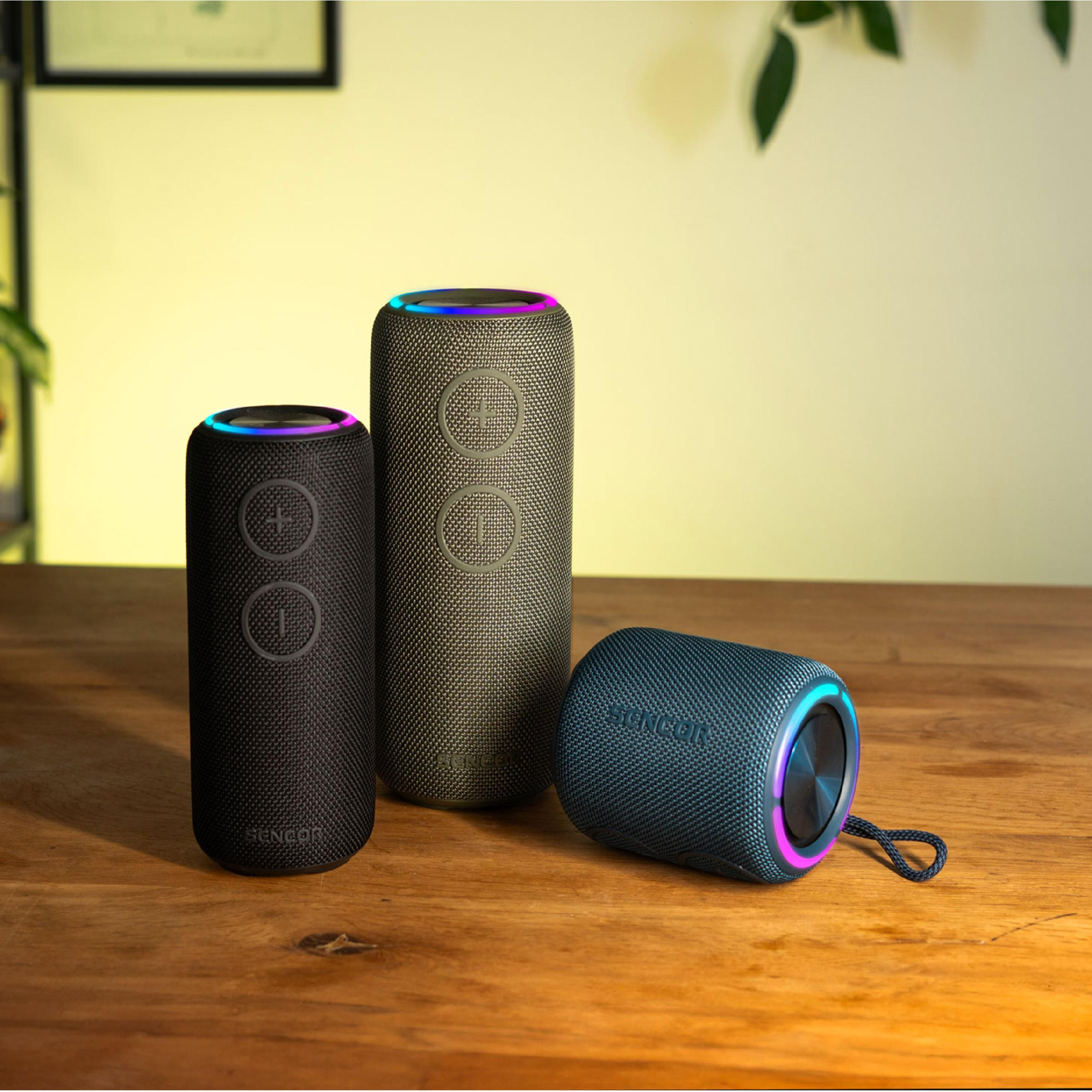 Trzy cylindryczne głośniki Bluetooth Sencor w kolorze czarnym, szarym i niebieskim z oświetleniem RGB stoją na drewnianym stole.