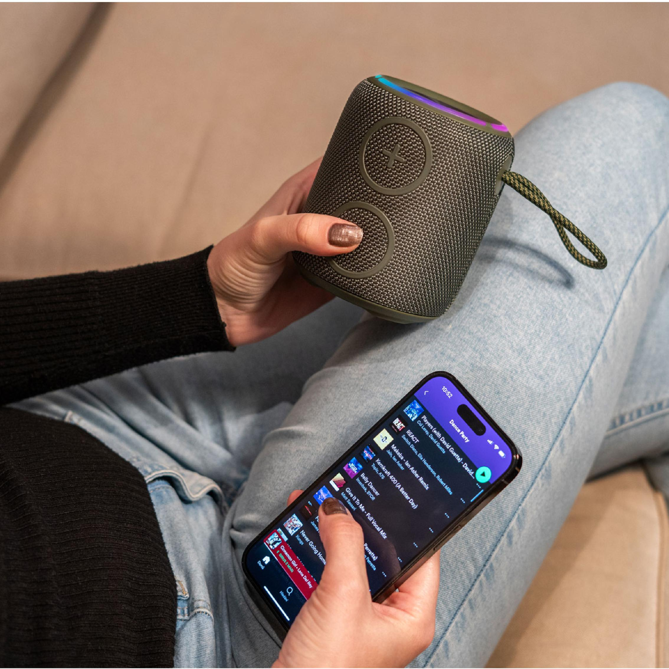 Osoba w dżinsach trzyma zielony głośnik Bluetooth i smartfon z aplikacją muzyczną.