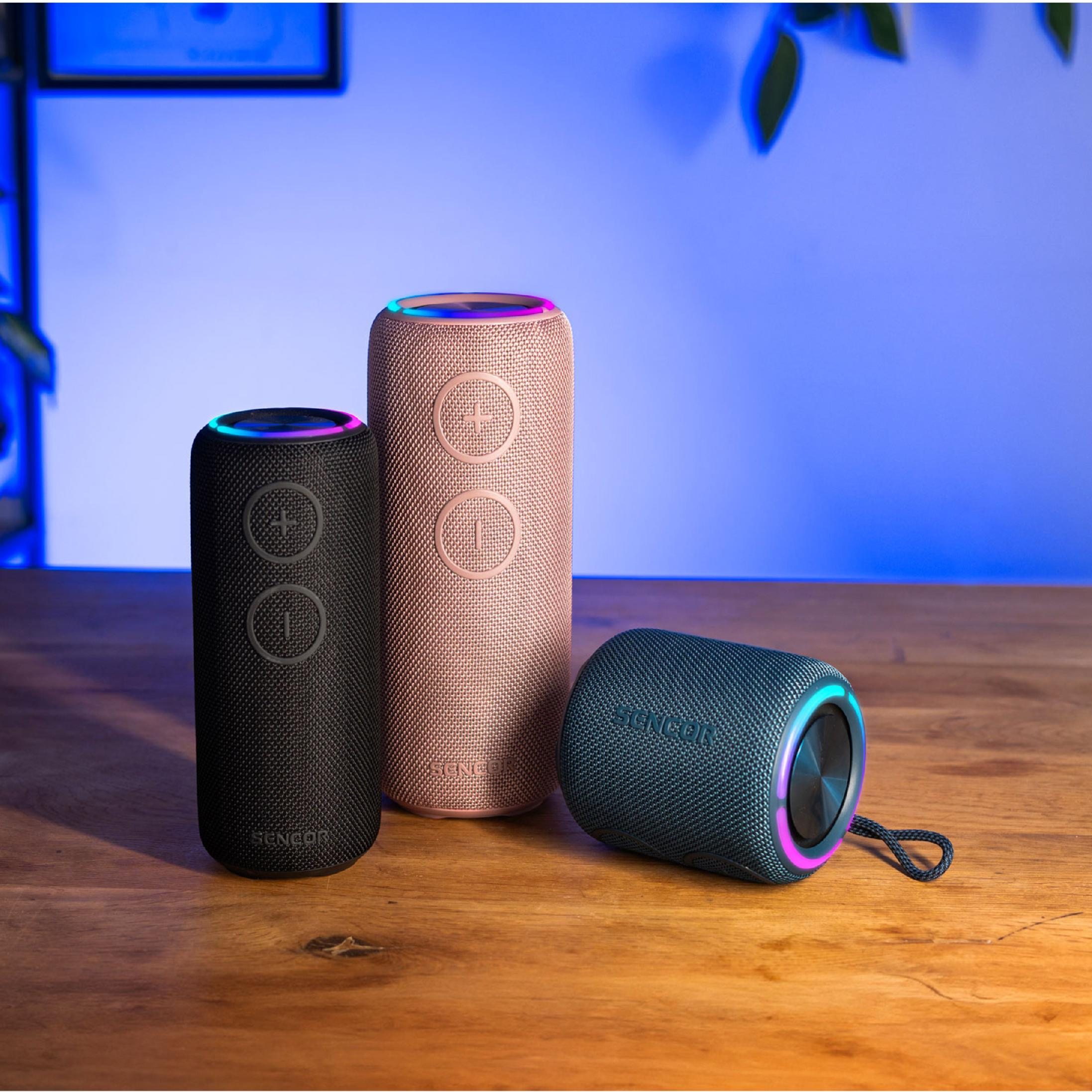 Trzy cylindryczne głośniki Bluetooth Sencor w kolorze czarnym, różowego złota i niebieskim stoją na drewnianym stole.