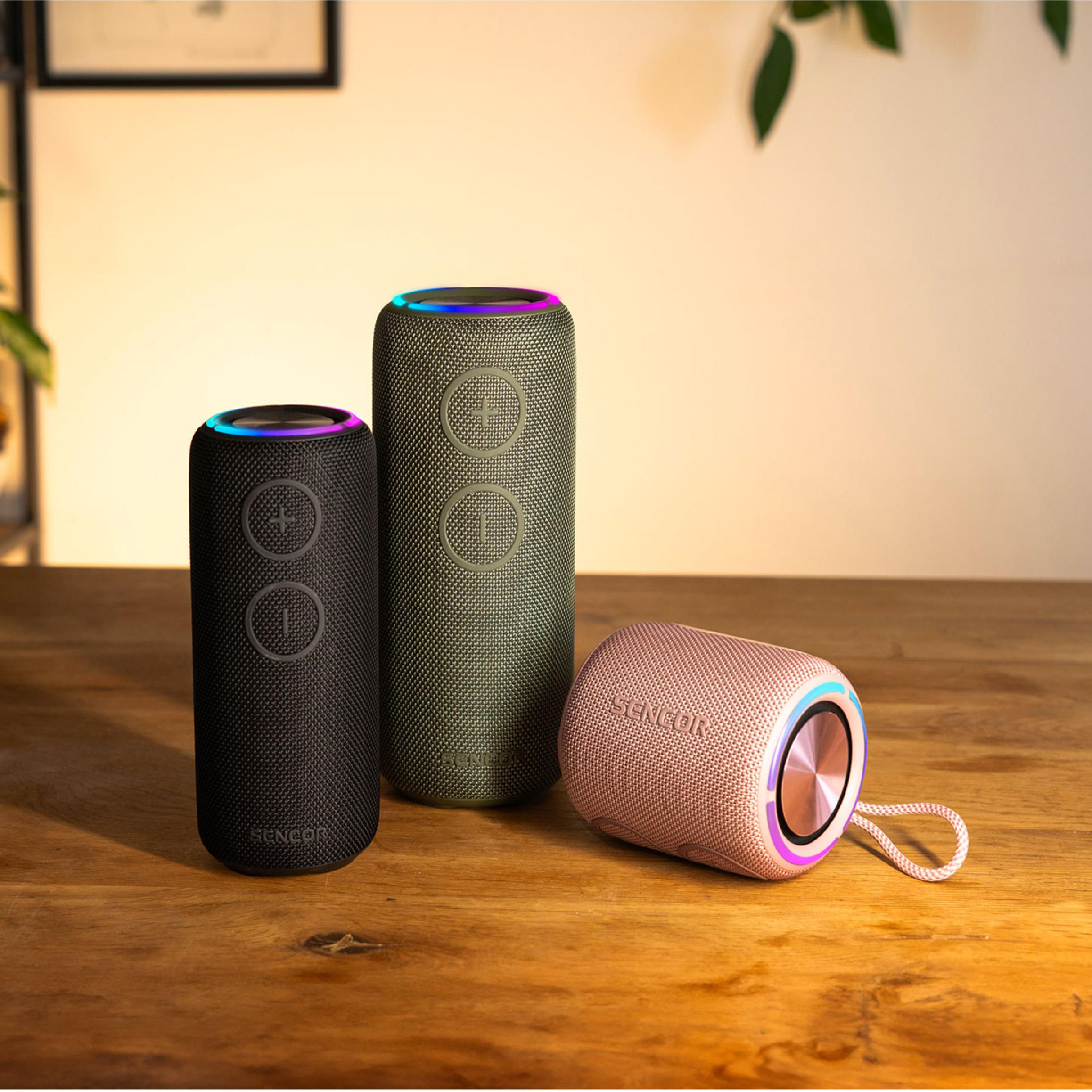 Trzy cylindryczne głośniki Bluetooth Sencor w kolorze czarnym, zielonym i różowym na drewnianym stole.