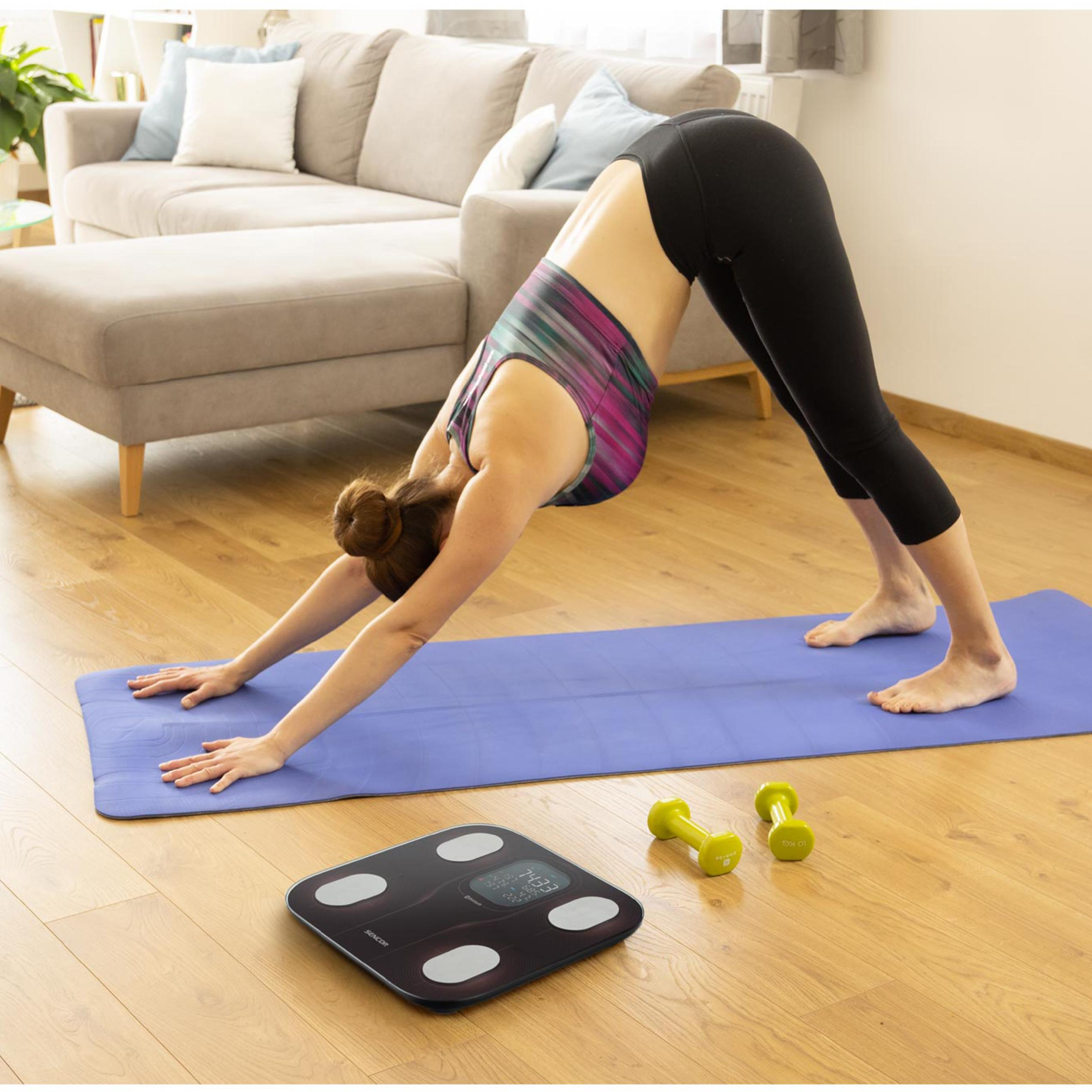 Eine Frau macht Yoga auf einer blauen Matte in einem Wohnzimmer; daneben liegen eine Waage und Hanteln.