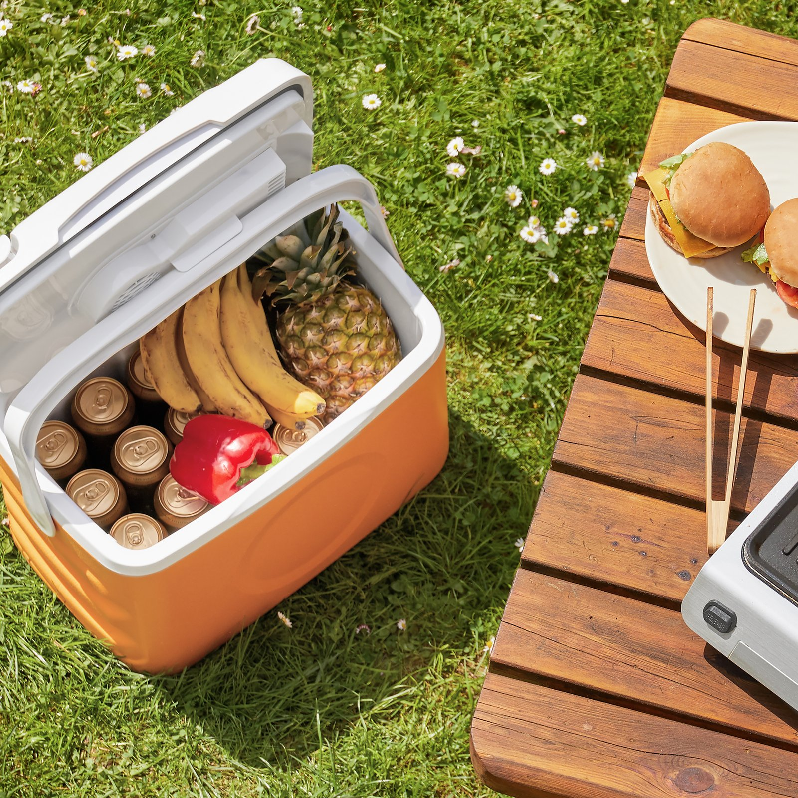 Eine offene orange Kühlbox mit Getränken und Früchten im Gras neben einem Holztisch mit Burgern.