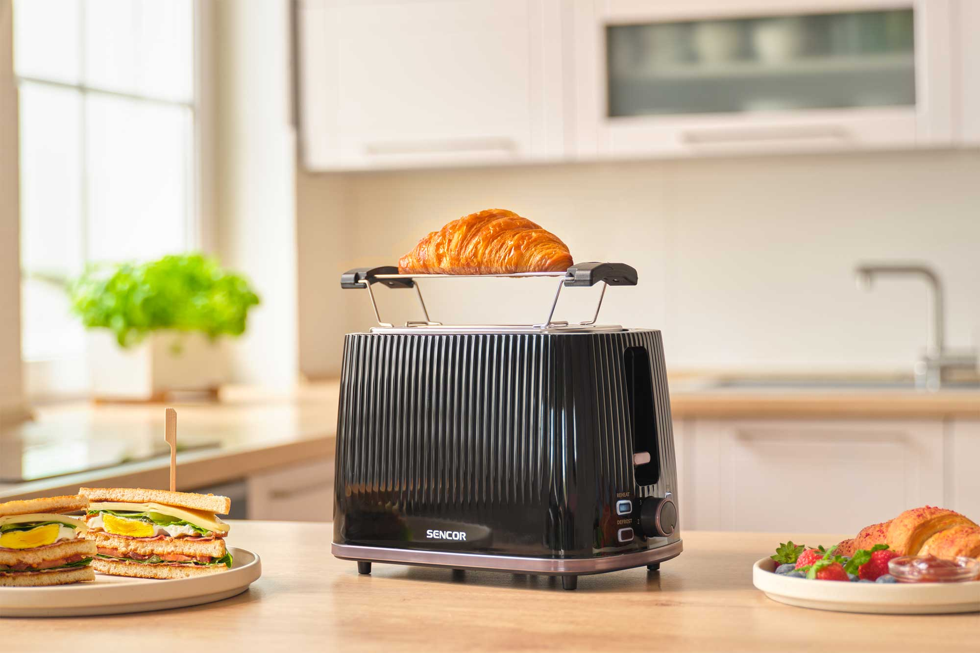 Czarny toster Sencor stoi na drewnianym blacie z rogalikiem podgrzewającym się na górze, w kuchni.