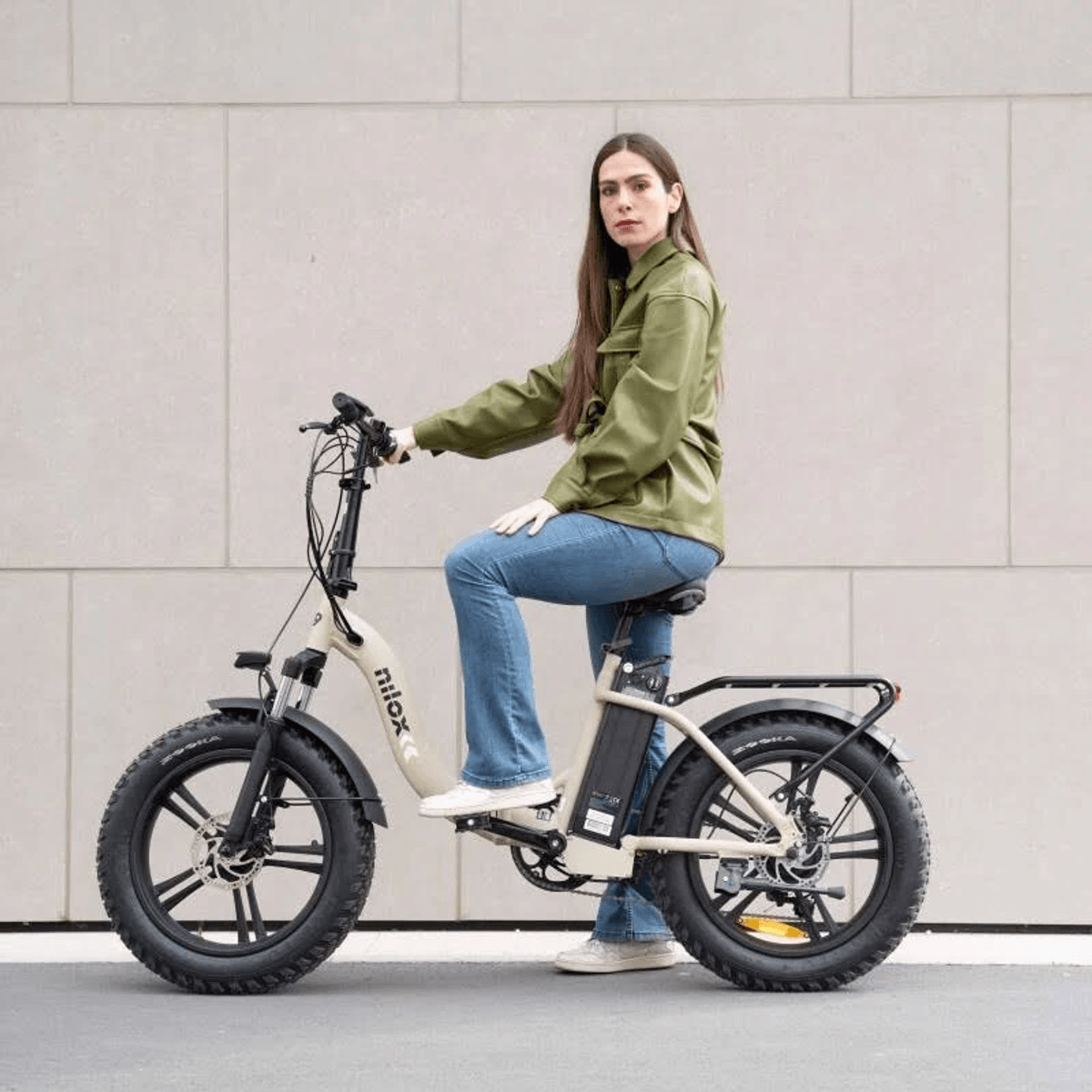 Una mujer con chaqueta verde y vaqueros se sienta en una bicicleta eléctrica de neumáticos anchos color crema.