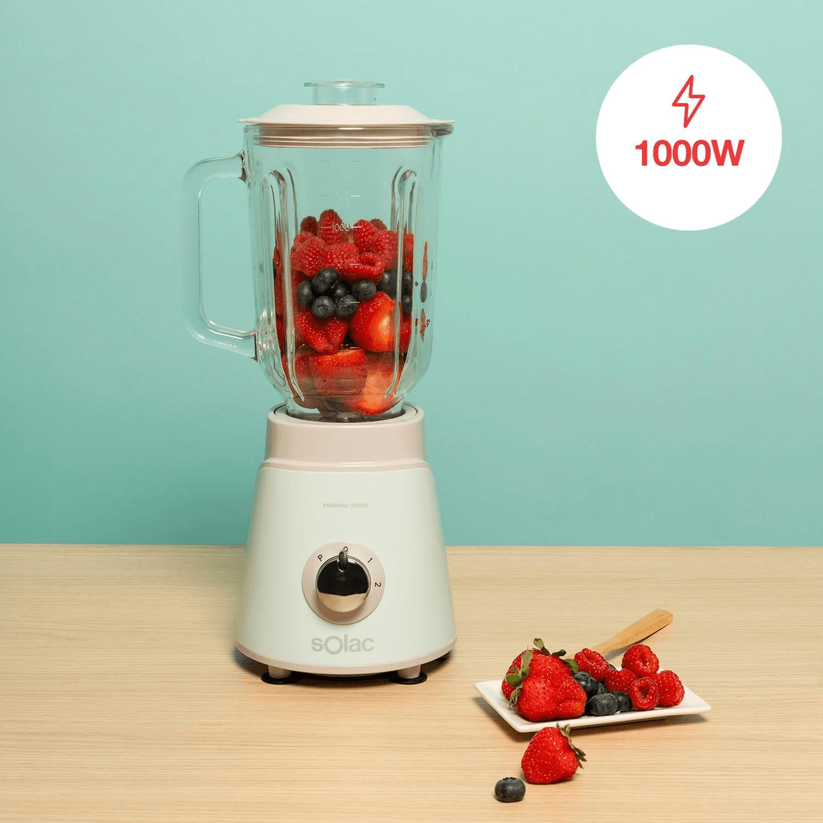 Una licuadora Solac blanca llena de fresas y arándanos sobre una mesa de madera con fondo verde azulado.