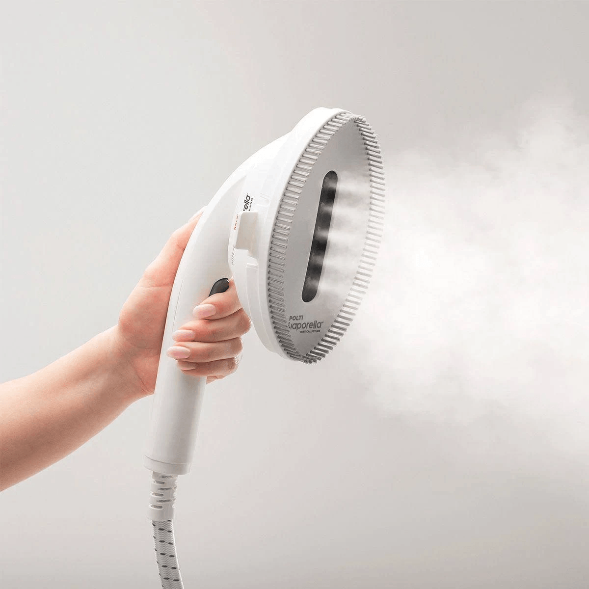 Una mano sostiene un vaporizador de prendas blanco que emite vapor sobre un fondo claro.