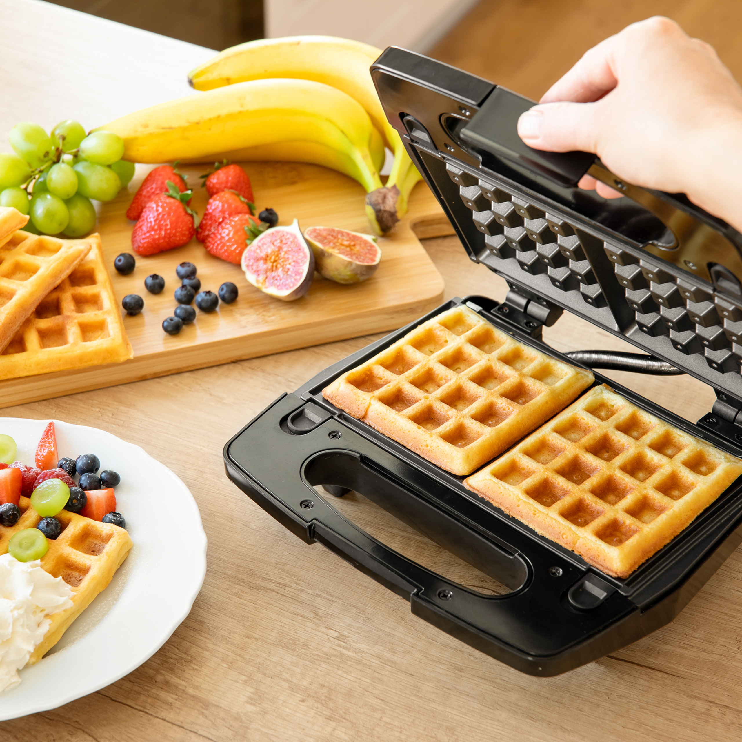 Hand öffnet Waffeleisen mit frisch gebackenen Waffeln, daneben Teller mit Früchten und Waffeln.