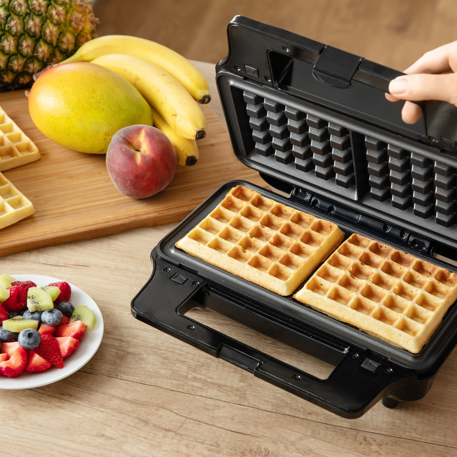 Ein Waffeleisen backt goldene Waffeln, daneben liegen frisches Obst wie Bananen, Beeren und eine Mango.