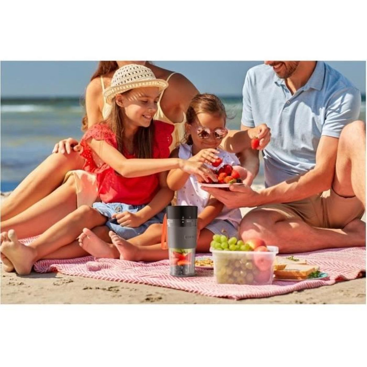 Szczęśliwa rodzina cieszy się piknikiem na plaży na czerwonym kocu, ze świeżymi owocami i przenośnym blenderem.
