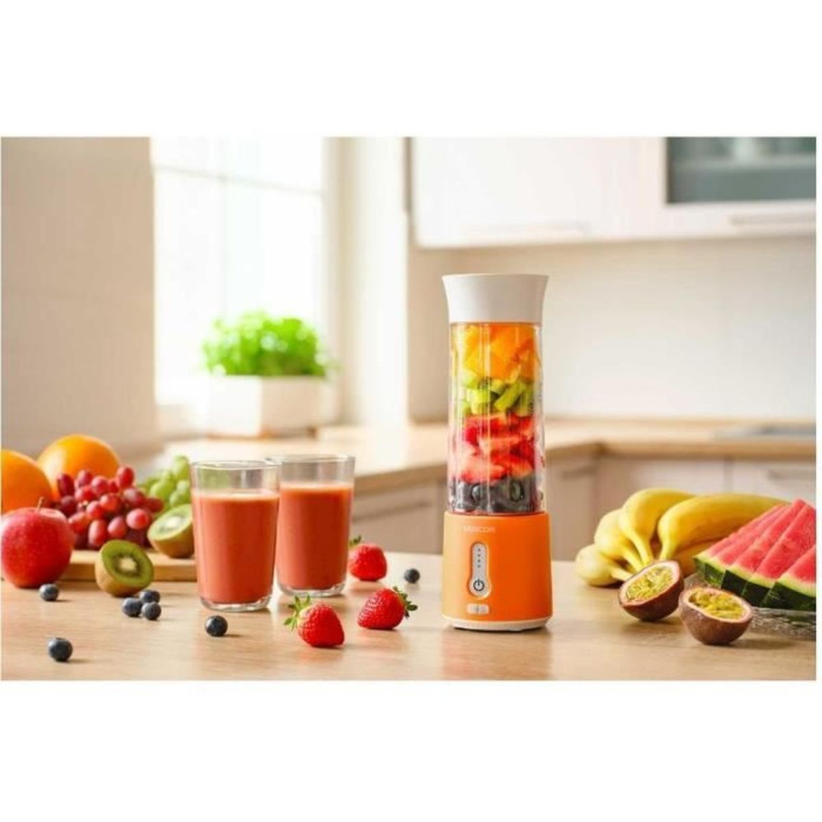 Pomarańczowy blender przenośny na blacie kuchennym, otoczony świeżymi owocami i dwoma szklankami soku.