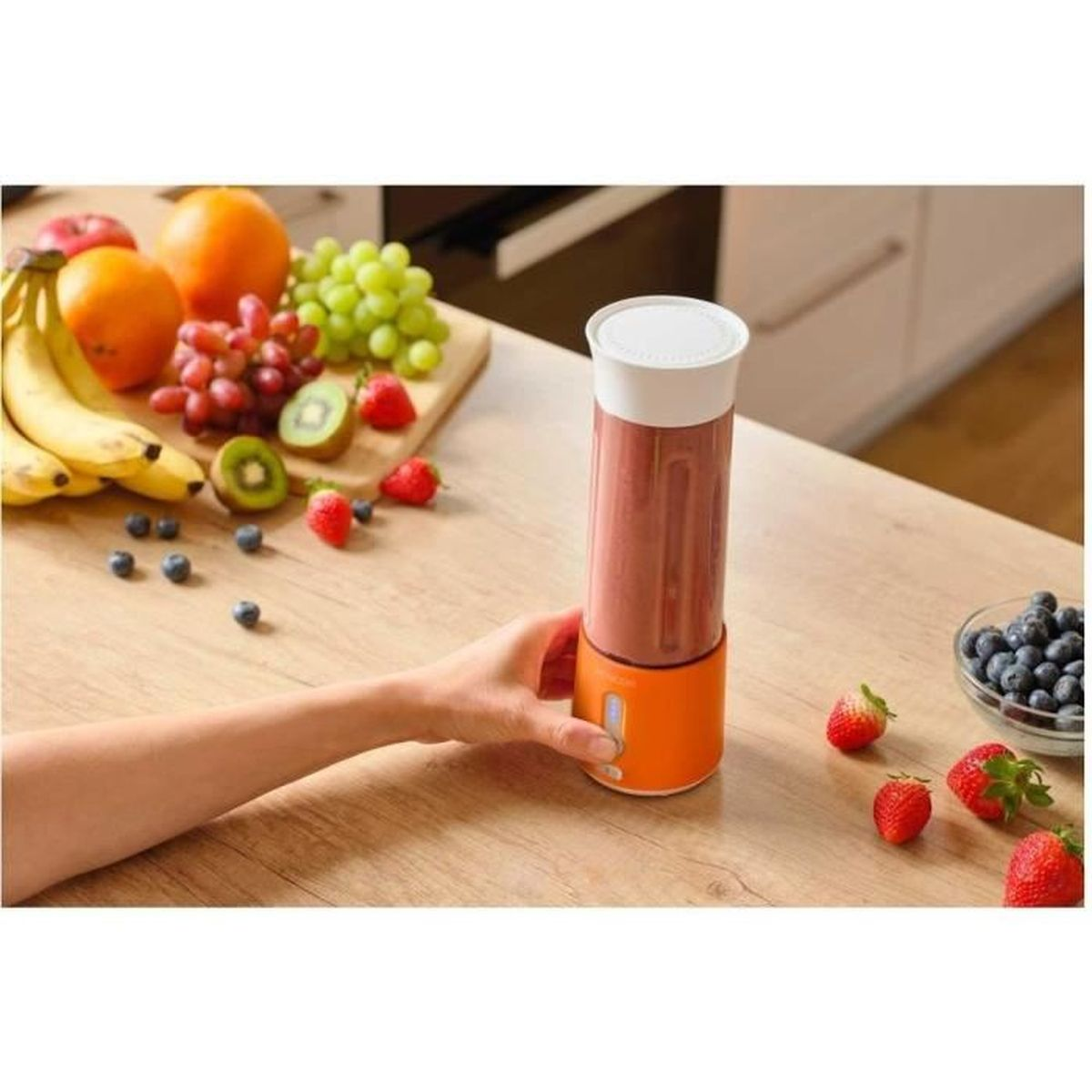 Ręka obsługuje pomarańczowy blender przenośny na drewnianym stole, w otoczeniu świeżych owoców.