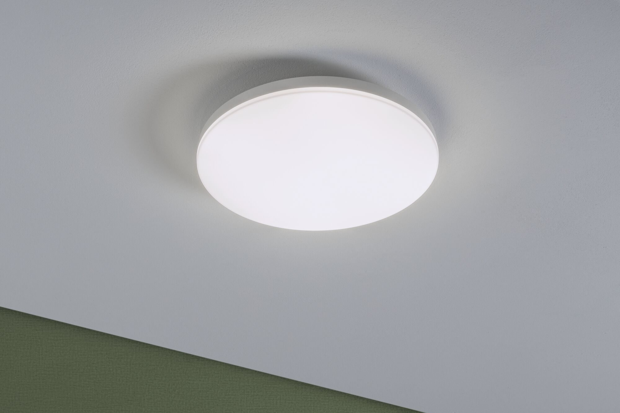 Eine runde, weiße Deckenlampe leuchtet an einer weißen Decke mit einem grünen Wandstreifen.