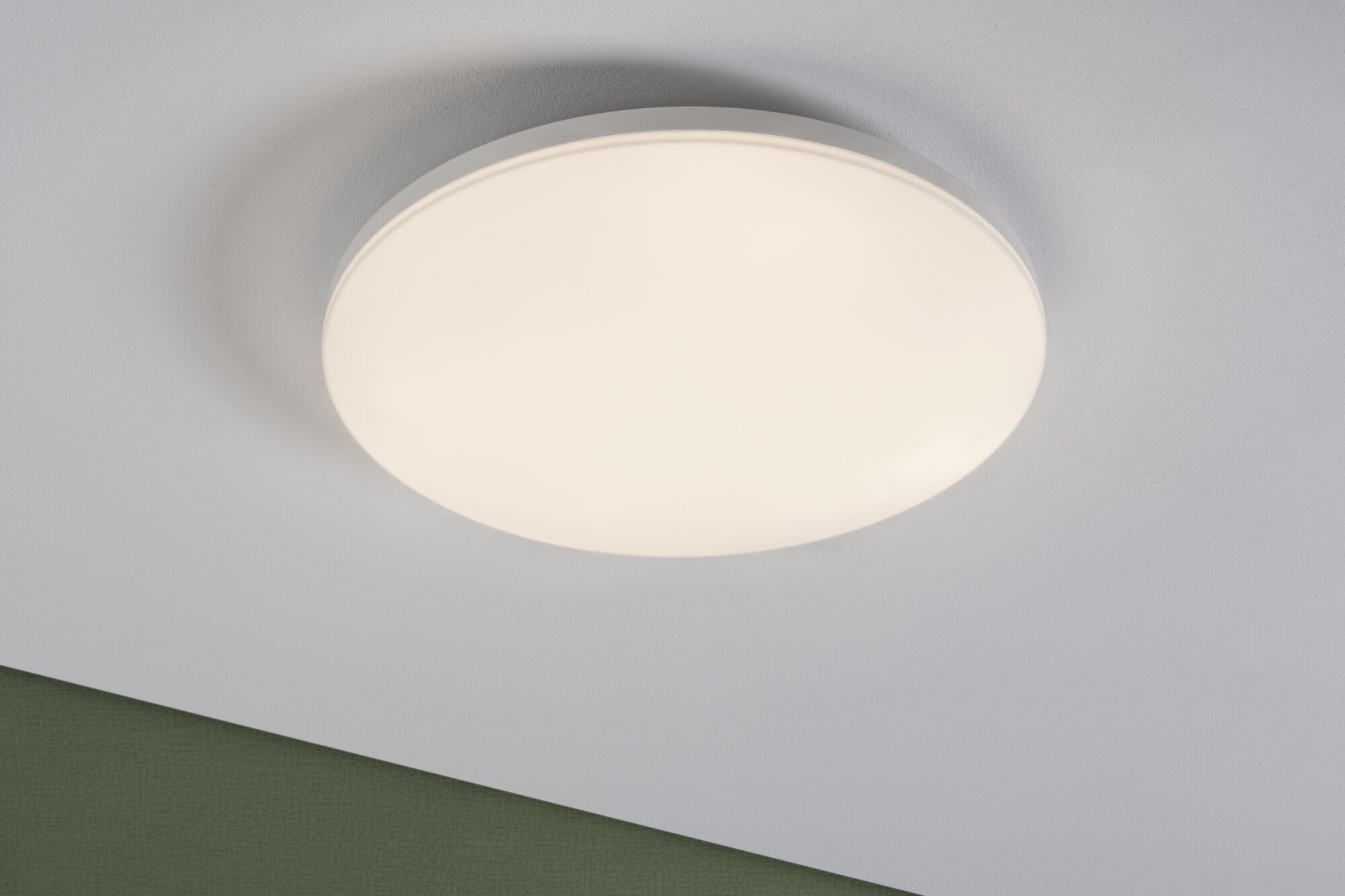 Eine runde, weiße LED-Deckenleuchte an einer weißen Decke mit einem grünen Wandabschnitt unten.