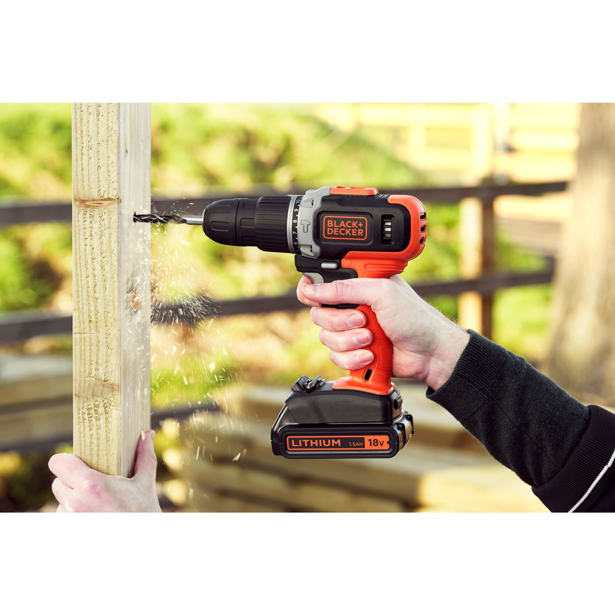 Eine Hand bohrt mit einer orangen und schwarzen Akkumaschine in einen Holzbalken, Späne fliegen.