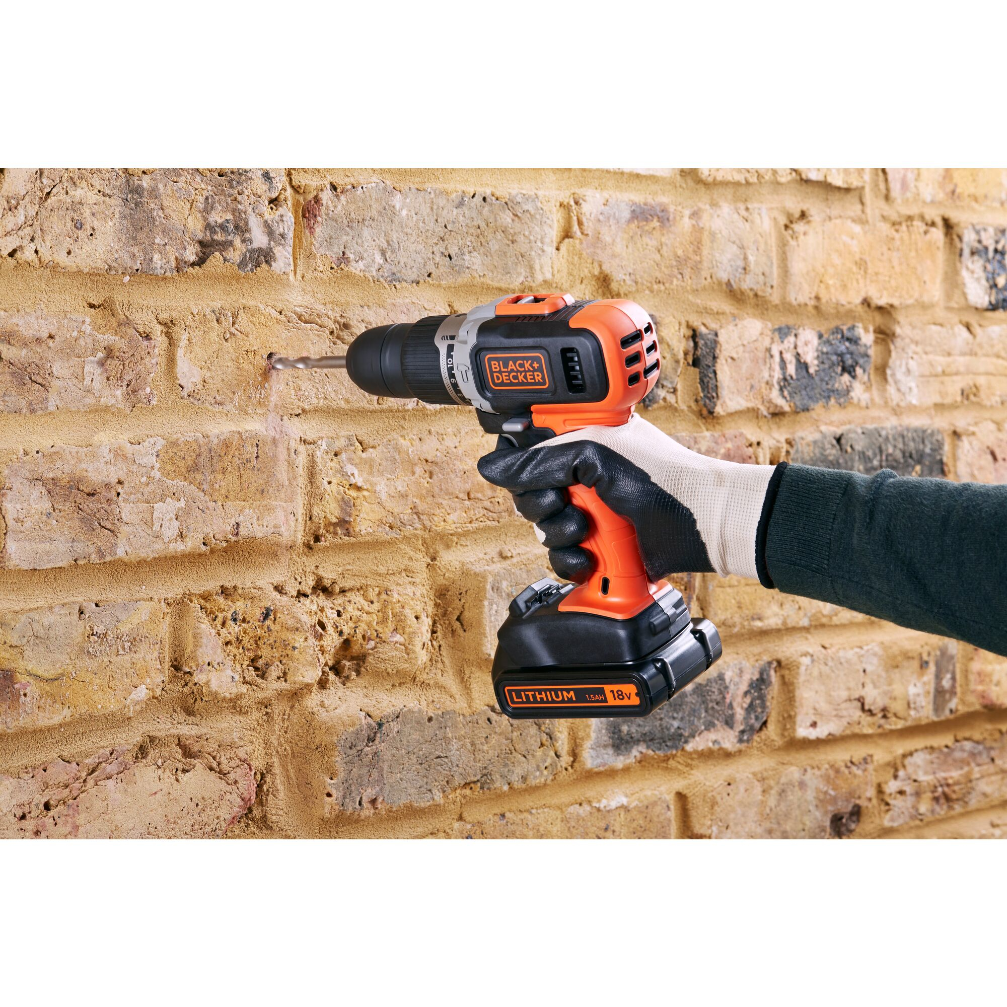 Eine Hand mit Handschuh bohrt ein Loch in eine gelbe Ziegelwand mit einer orange-schwarzen Akkubohrmaschine.