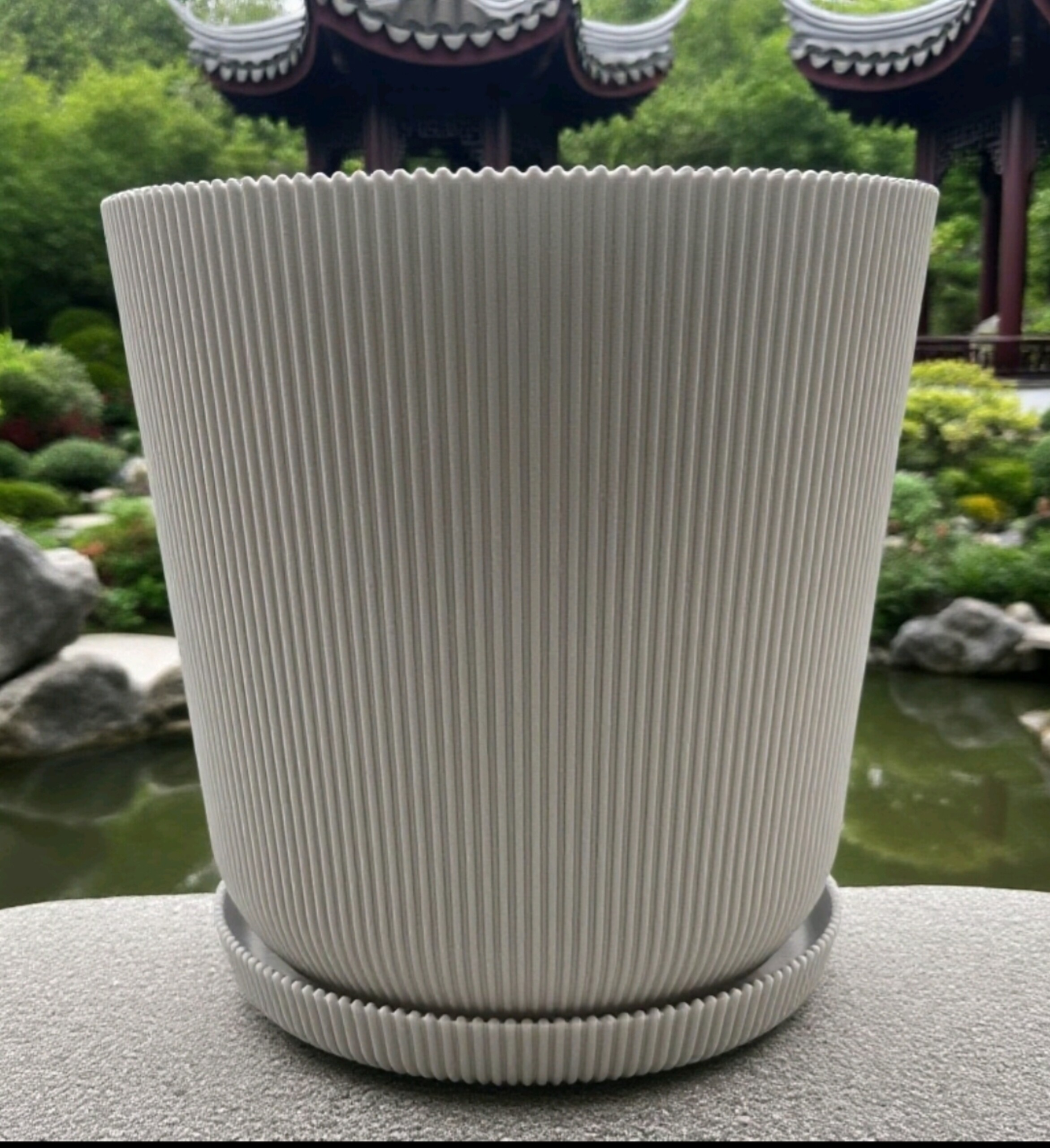 Ein weißer, gerippter Blumentopf mit Untersetzer auf einer Steinplatte im Garten vor einem Teich und Pavillon.