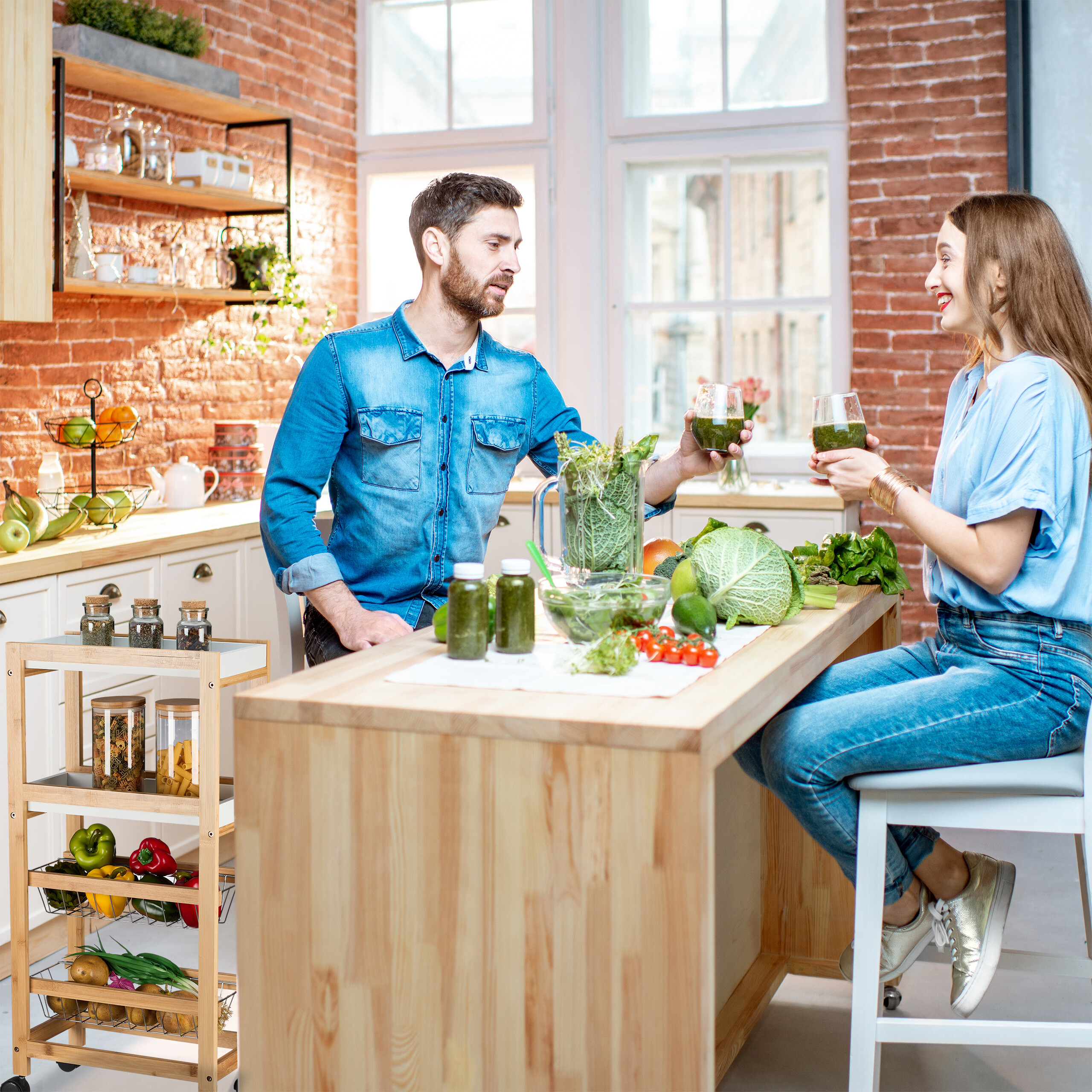 Ein Mann und eine Frau in einer rustikalen Küche stoßen mit grünen Smoothies an, auf einer Theke voller Gemüse.