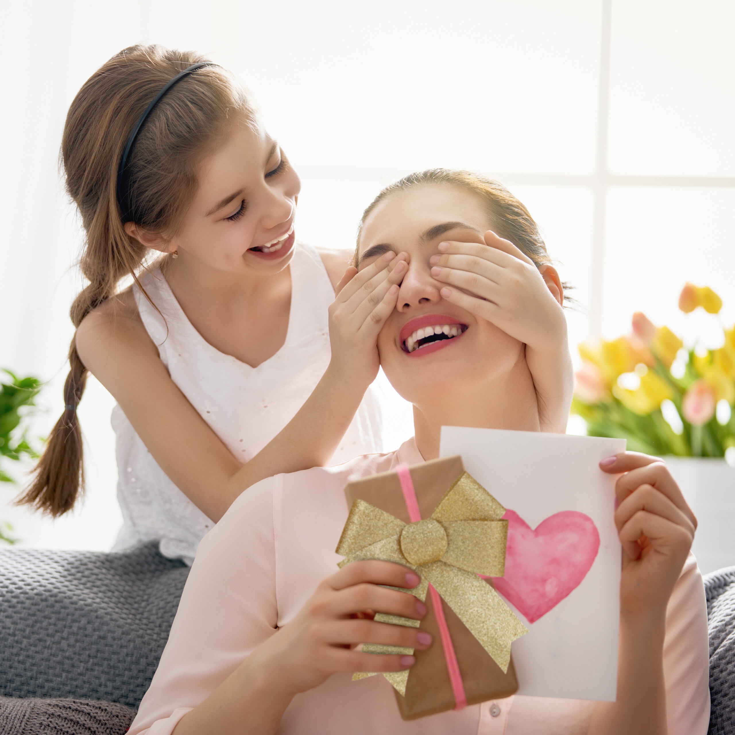 Ein smiling Mädchen bedeckt die Augen ihrer Mutter, die ein Geschenk und eine Karte hält.
