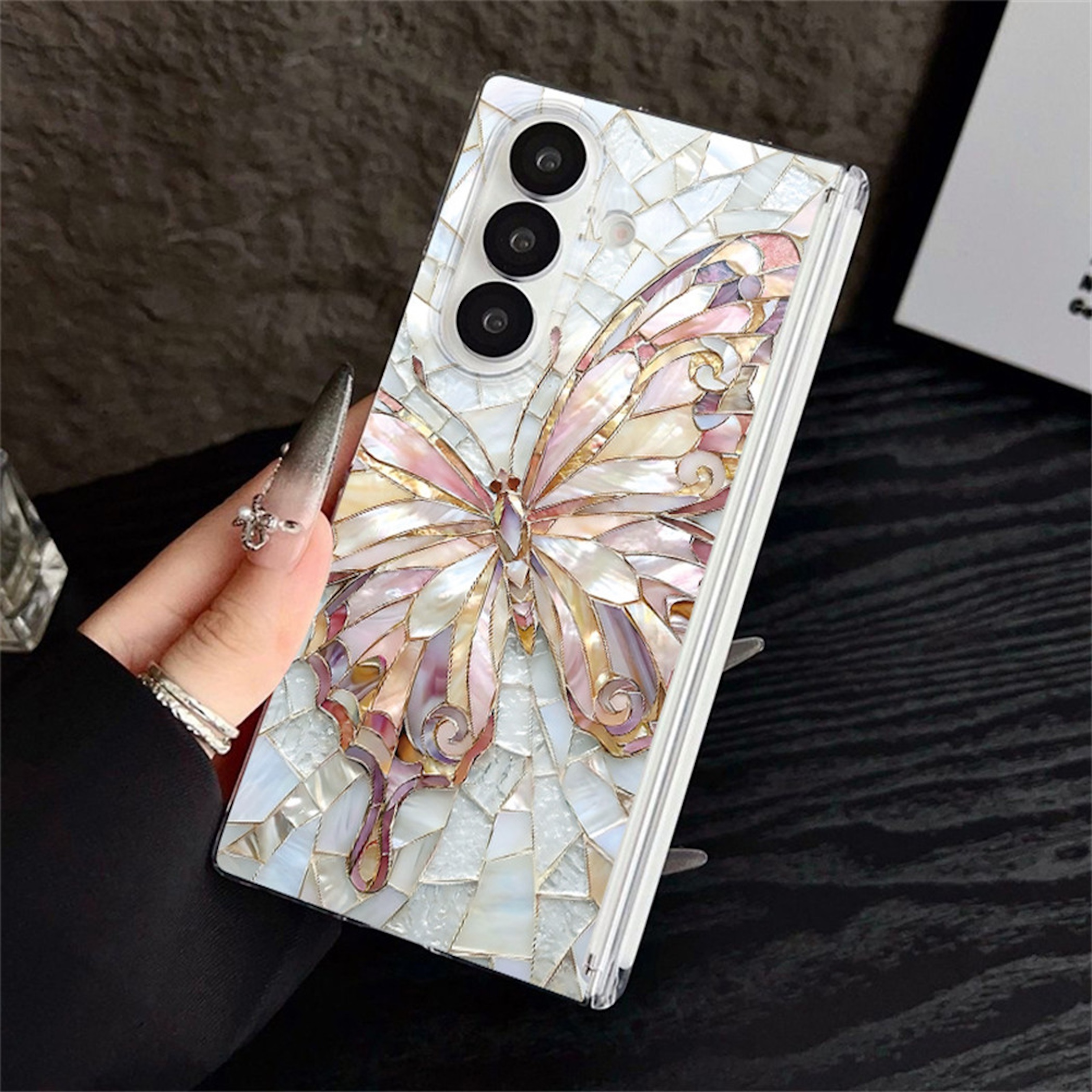 Eine Hand hält ein Smartphone mit einer Perlmutt-Mosaik-Schmetterlingshülle auf einer dunklen Oberfläche.