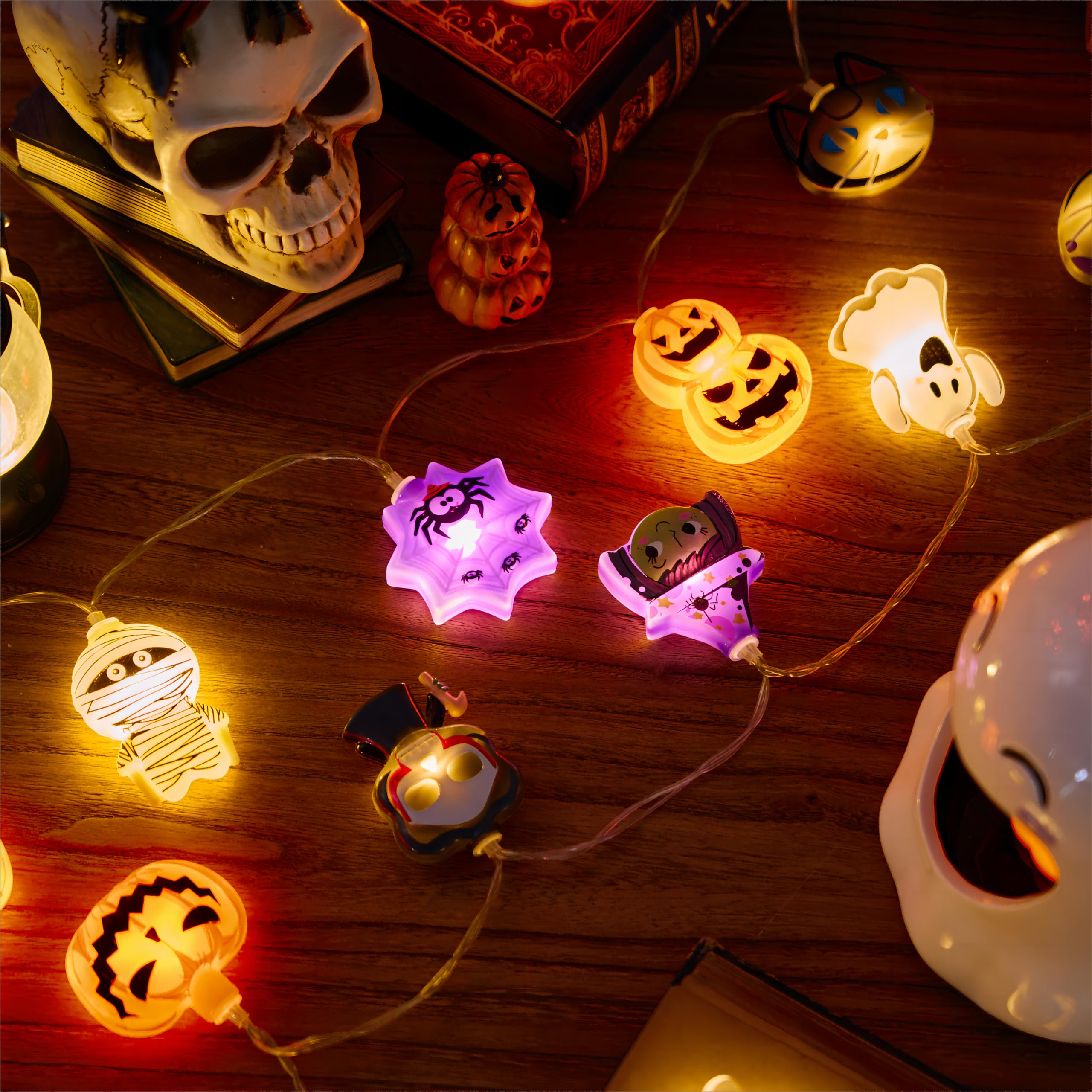 Eine Halloween-Dekoration mit einer Lichterkette, einem Totenkopf, kleinen Kürbissen und Geisterfiguren auf Holz.