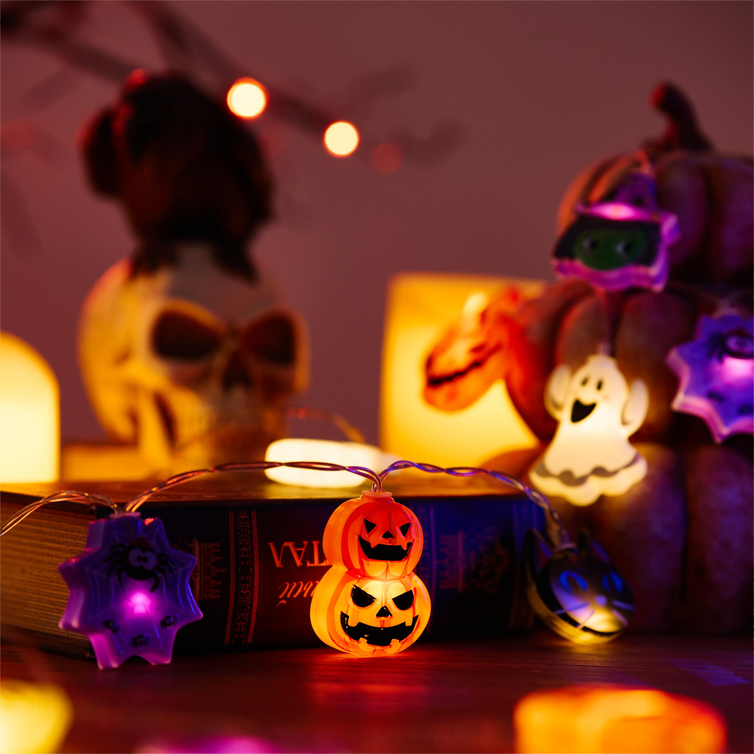 Halloween-Dekoration mit Kürbislichterkette, Buch, Totenkopf und flackernden Kerzen im Hintergrund.