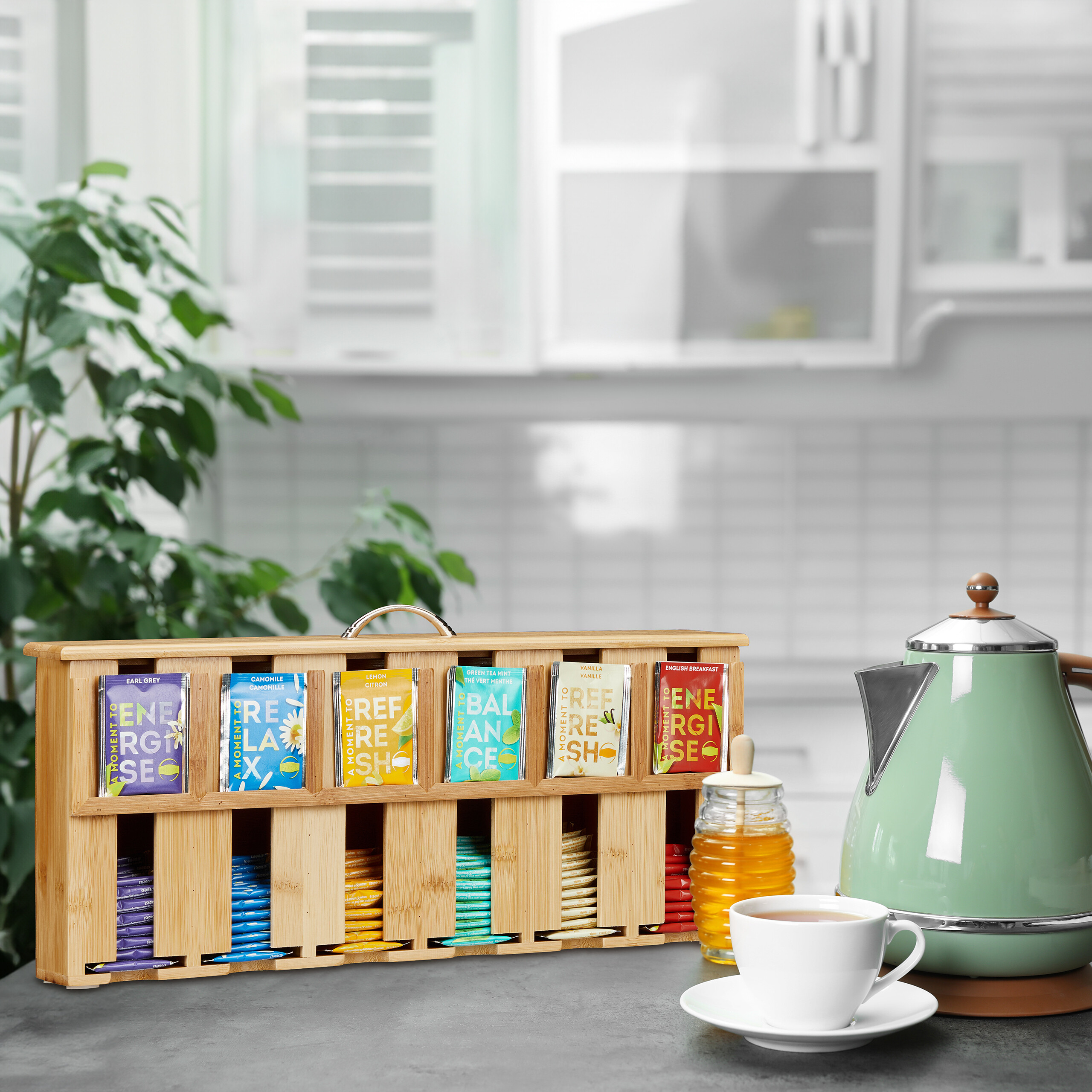 Ein Holz-Teebeutelhalter, eine Teekanne, eine Tasse Tee und Honig stehen auf einer Küchentheke.