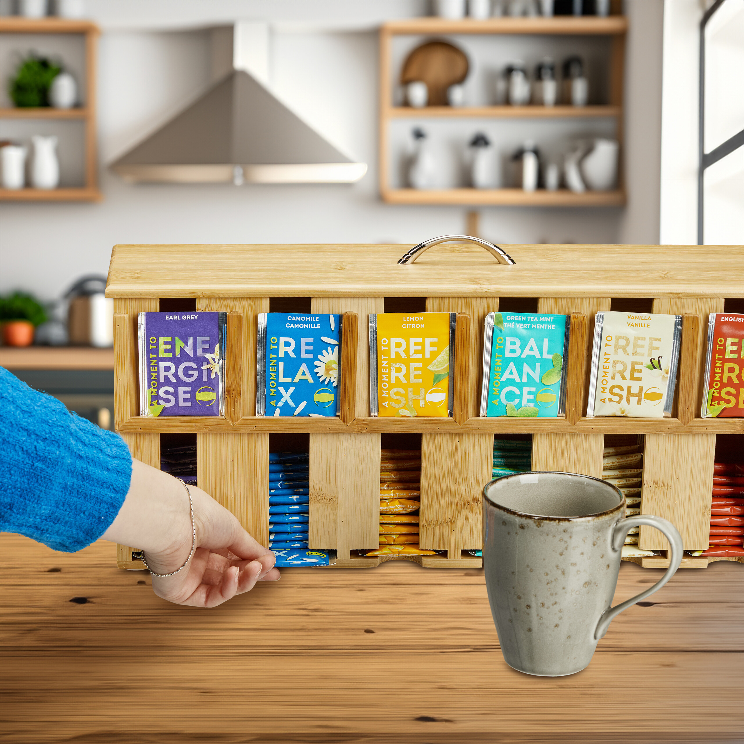 Hand greift in hölzerne Tee-Organizer-Box mit bunten Tütchen auf Küchentisch, daneben graue Tasse.