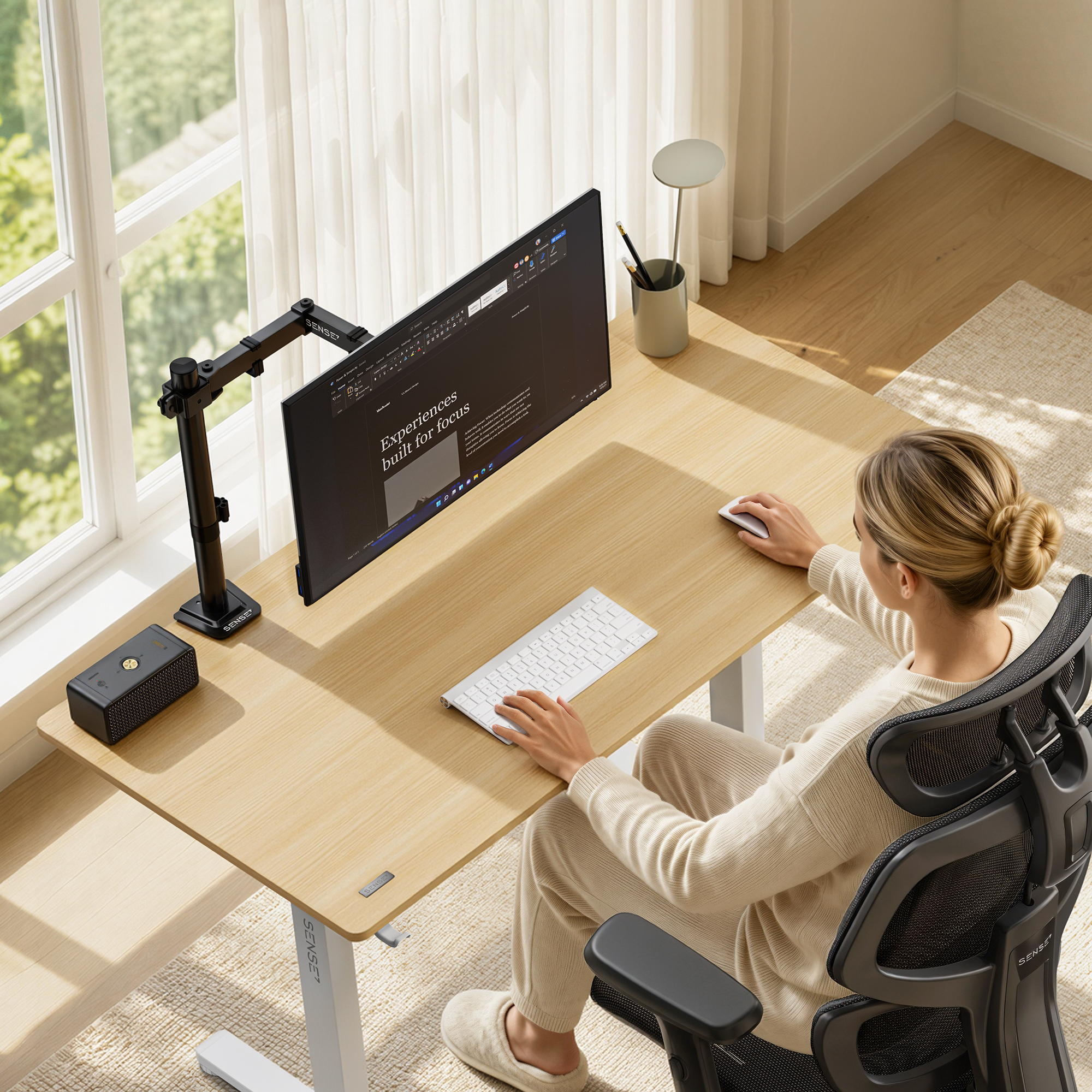 Eine Frau arbeitet an einem Holztisch mit Monitor, Tastatur, Maus und Stuhl an einem sonnigen Fenster.