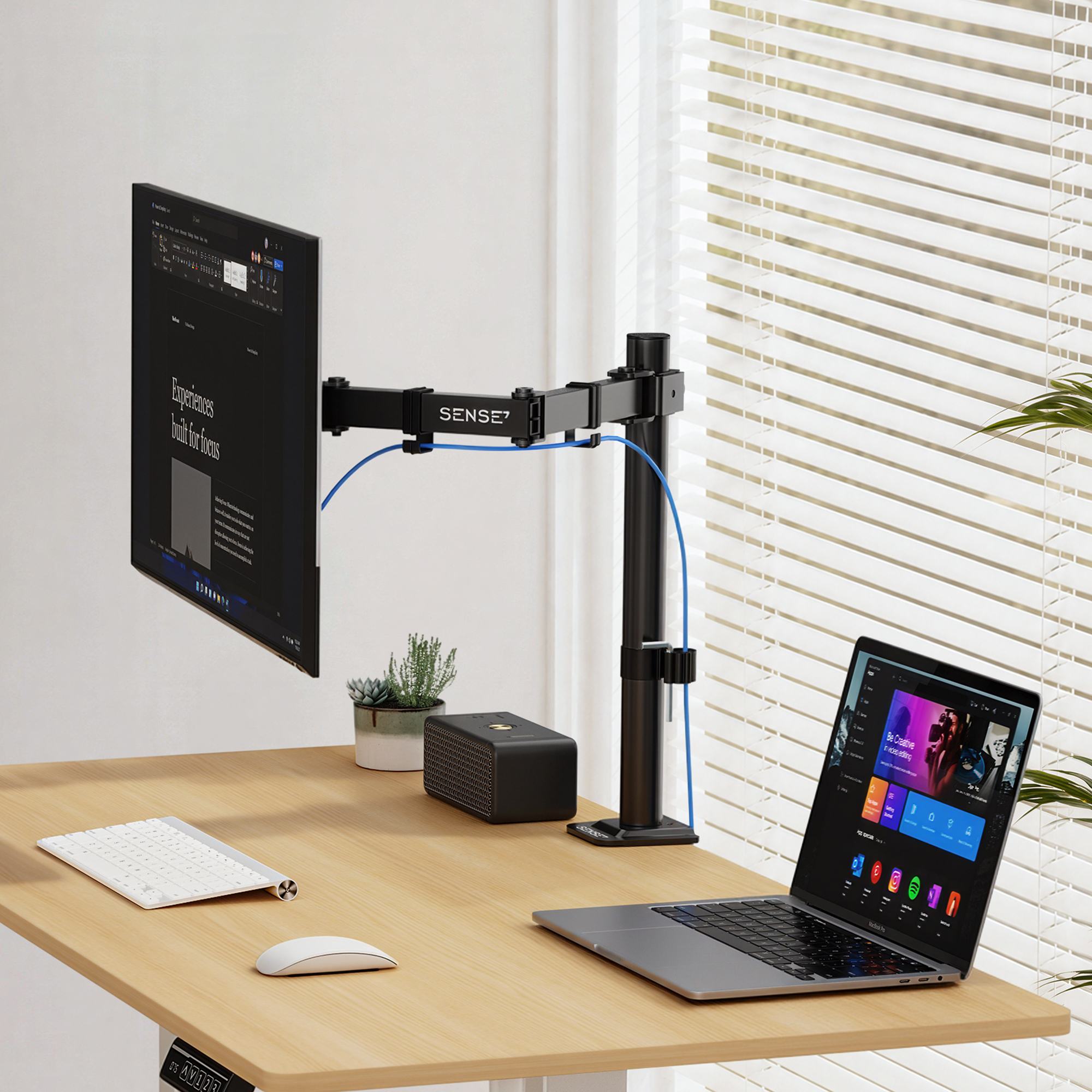Ein Schreibtisch mit Monitorarm, Laptop, weißer Tastatur, Maus und Lautsprecher vor einem Fenster mit Jalousien.