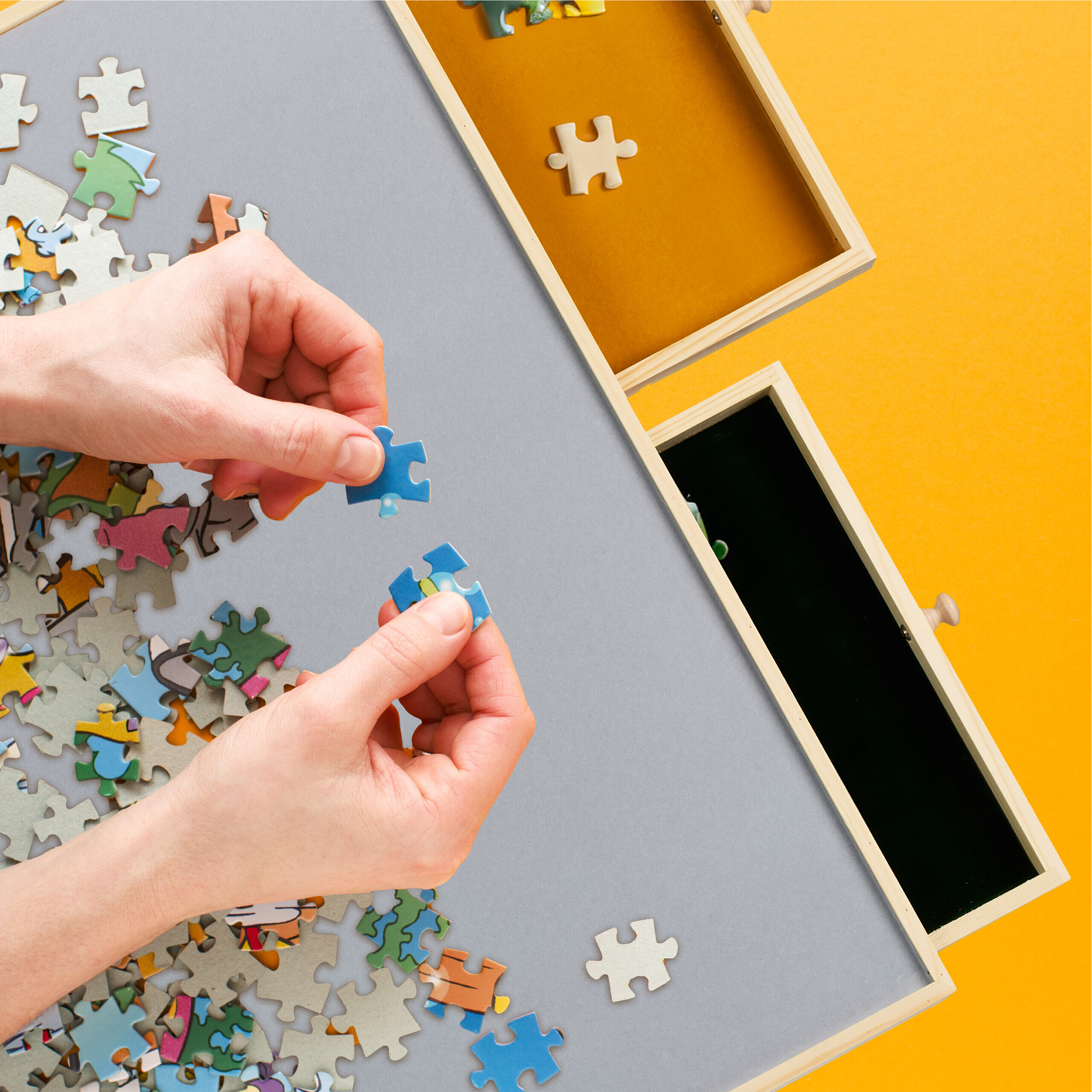 Hände setzen blaue Puzzleteile auf ein graues Brett, mit hölzernen Sortierkästen und verstreuten Teilen.