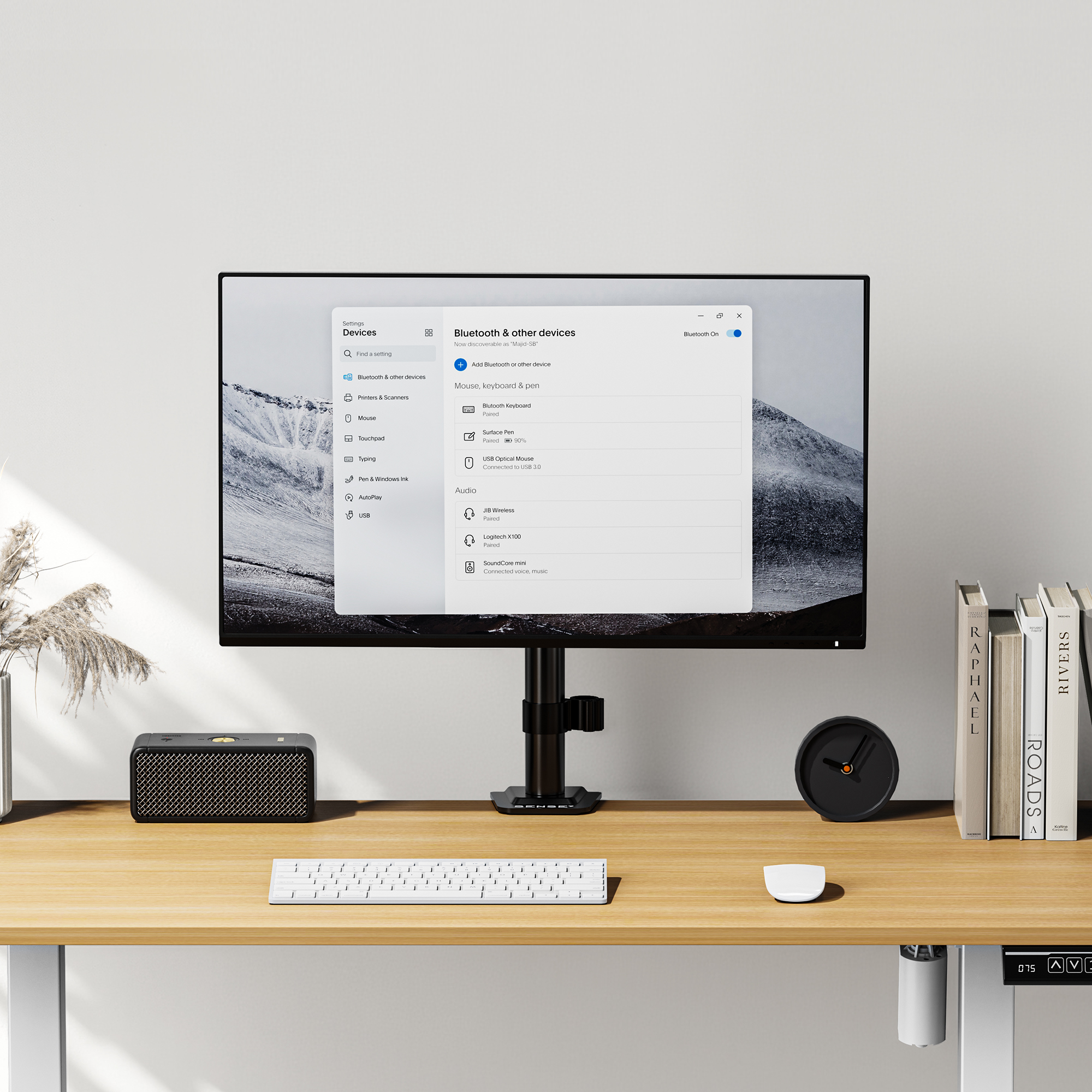 Ein minimalistischer Arbeitsbereich mit einem Monitor, der Bluetootheinstellungen anzeigt, einer Tastatur und Büchern.