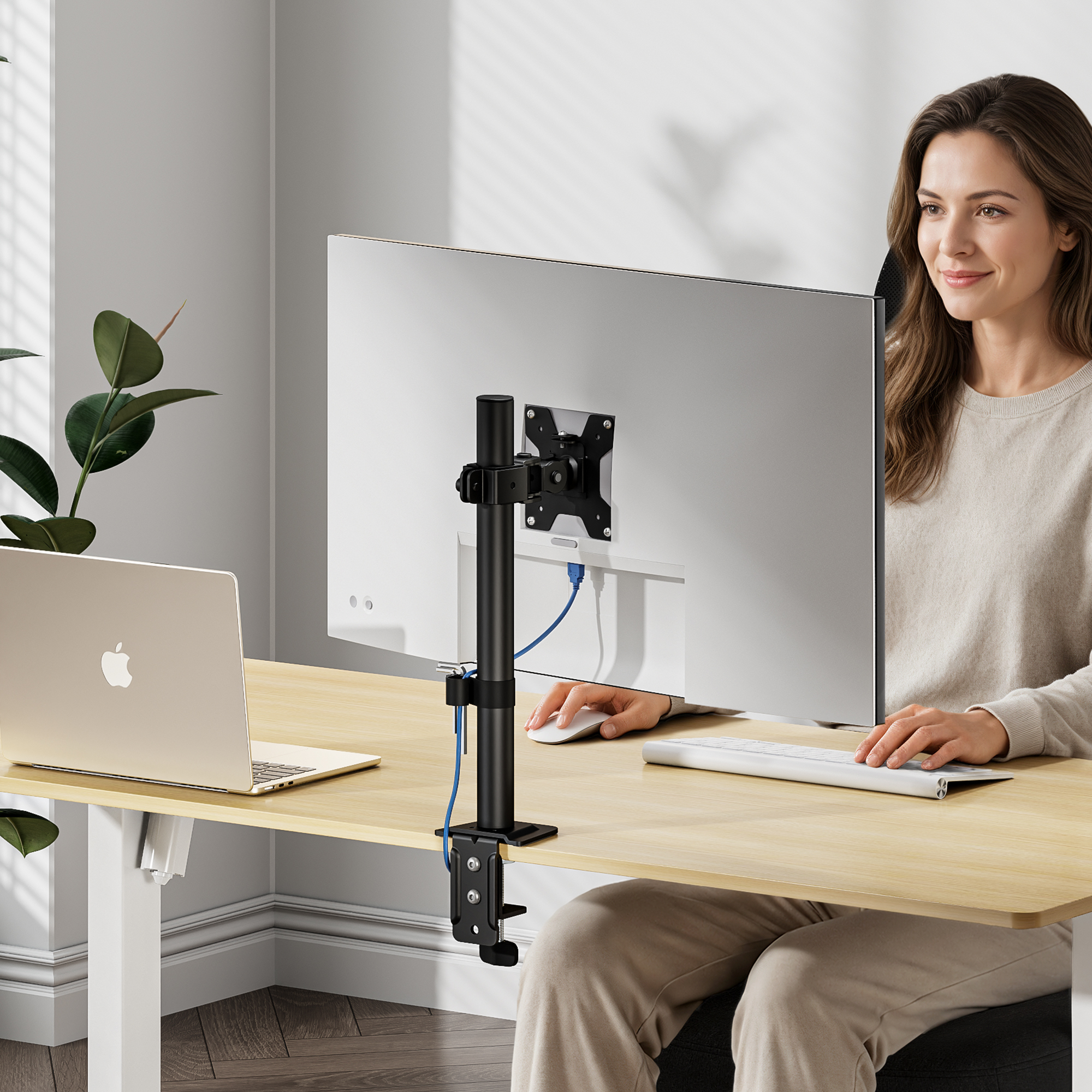 Eine Frau arbeitet an einem Holztisch mit einem Monitor auf schwarzem Ständer, einem Laptop und einer Pflanze.
