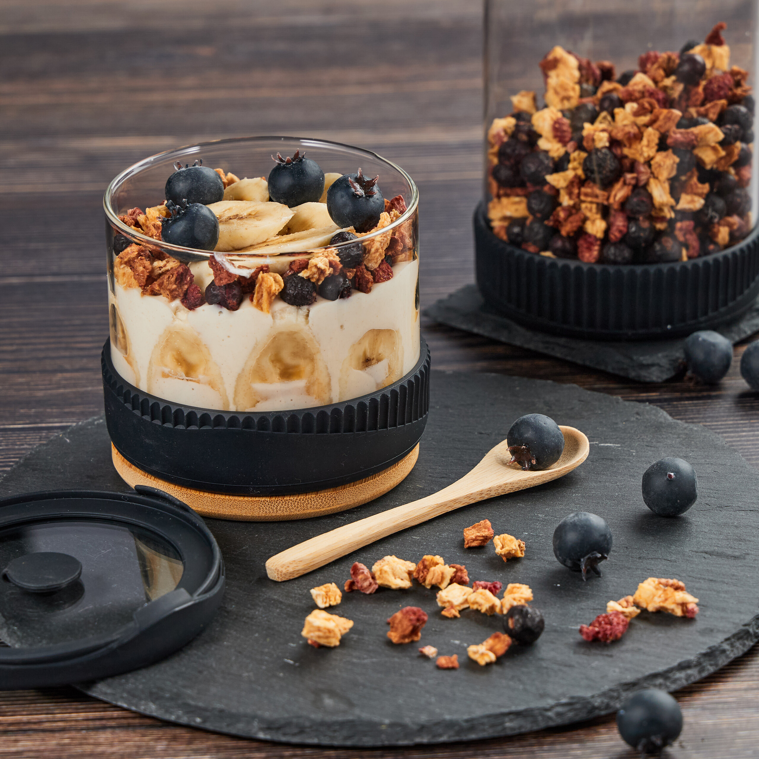 Ein Glas Joghurt mit Bananen, Blaubeeren und Müsli auf einer Schieferplatte, daneben lose Beeren und Getreide.