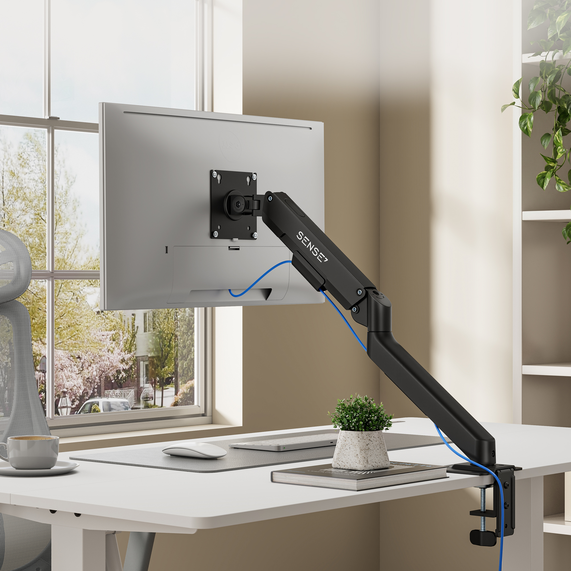 Ein weißer Monitor an einem schwarzen SENSE7-Arm auf einem Schreibtisch mit Pflanze und Fenster im Hintergrund.