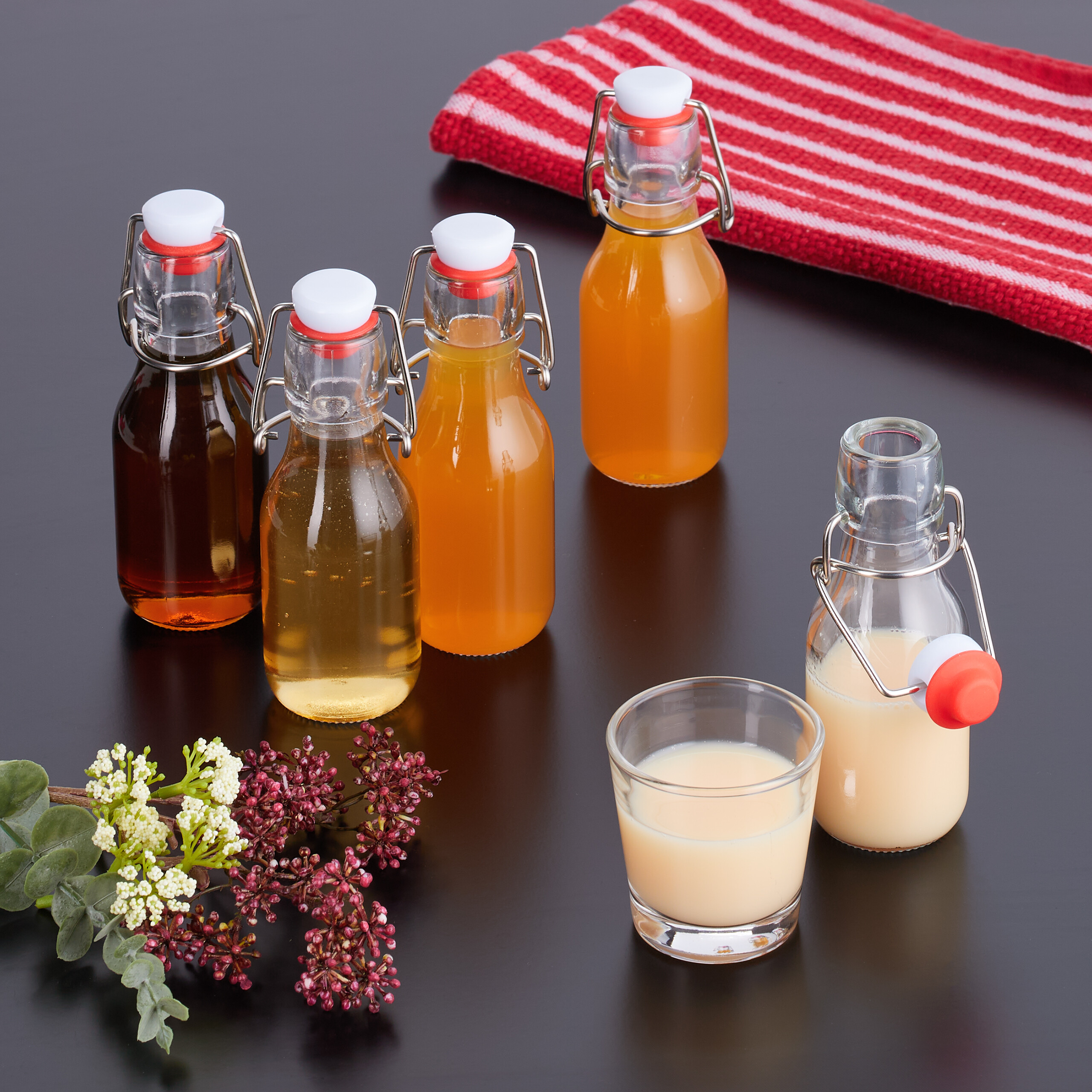 Glasflaschen mit verschiedenen Getränken, ein Glas Milch, Blumen und ein rot-weißes Tuch auf dunklem Tisch.