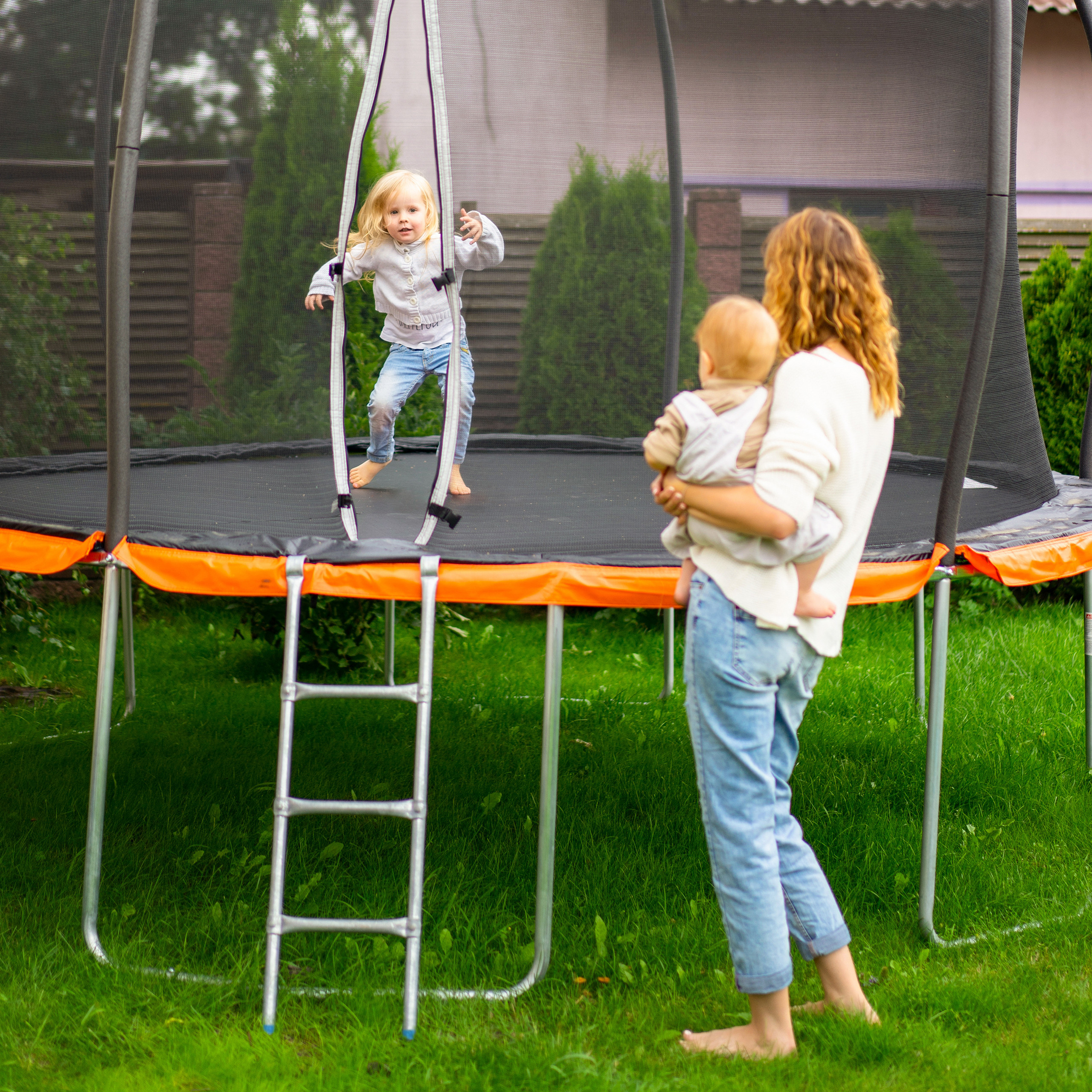 Eine Frau hält ein Baby im Arm, während ein Kind auf dem Trampolin springt.