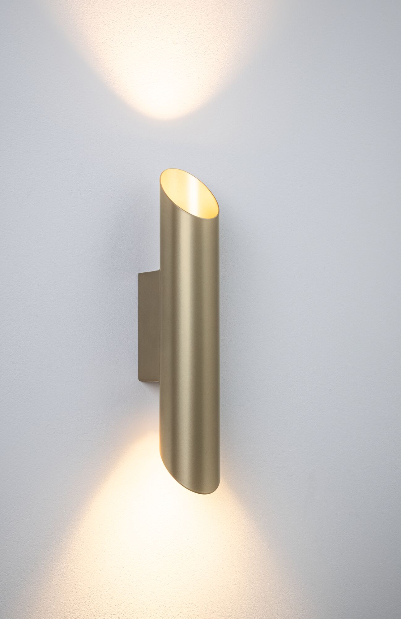 Eine goldfarbene zylindrische Wandleuchte leuchtet nach oben und unten an einer grauen Wand.